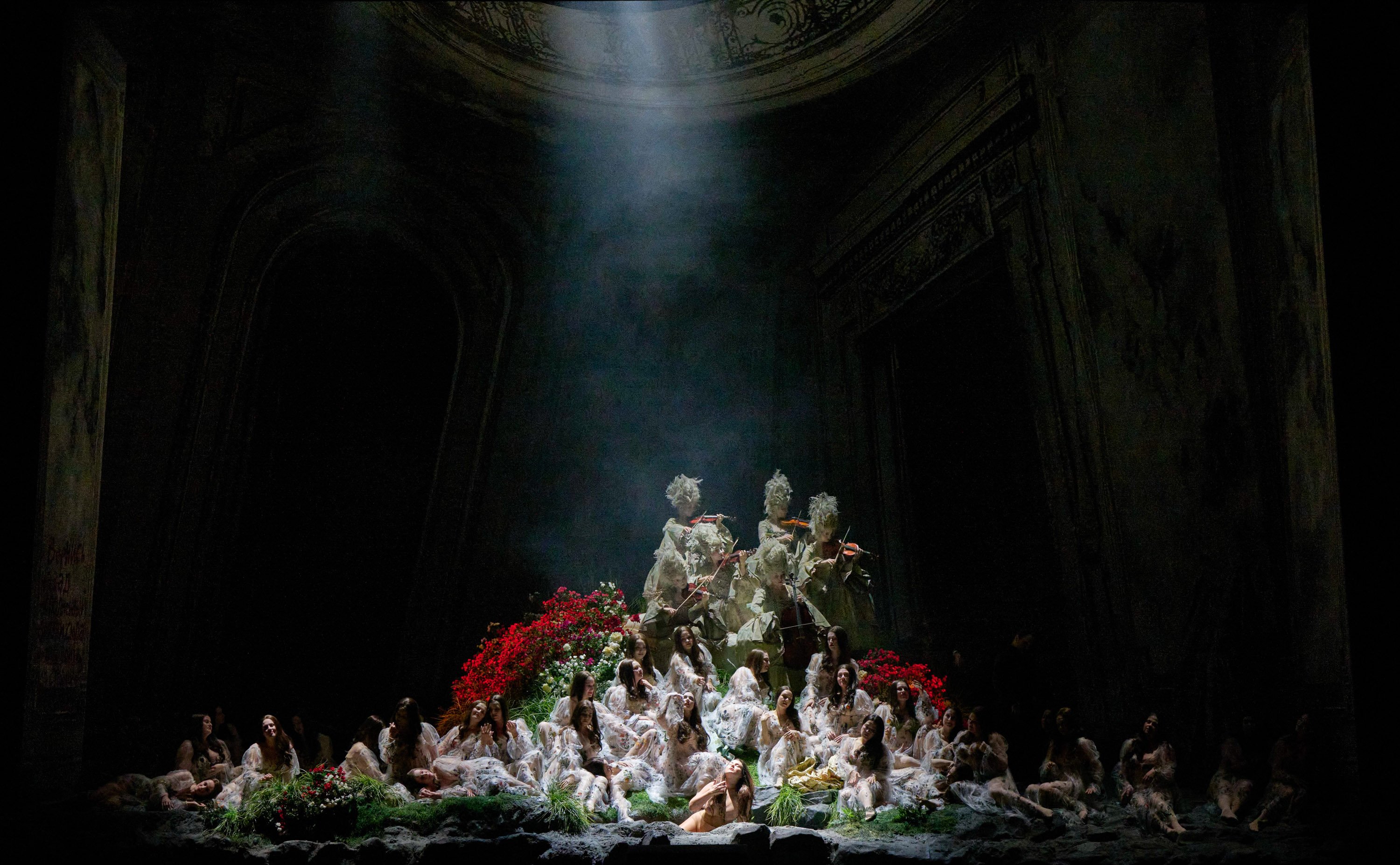 In geheimnisvollem Bühnenlicht sitzt eine Frauen-Gruppe auf einem blumenumrahmten Podest