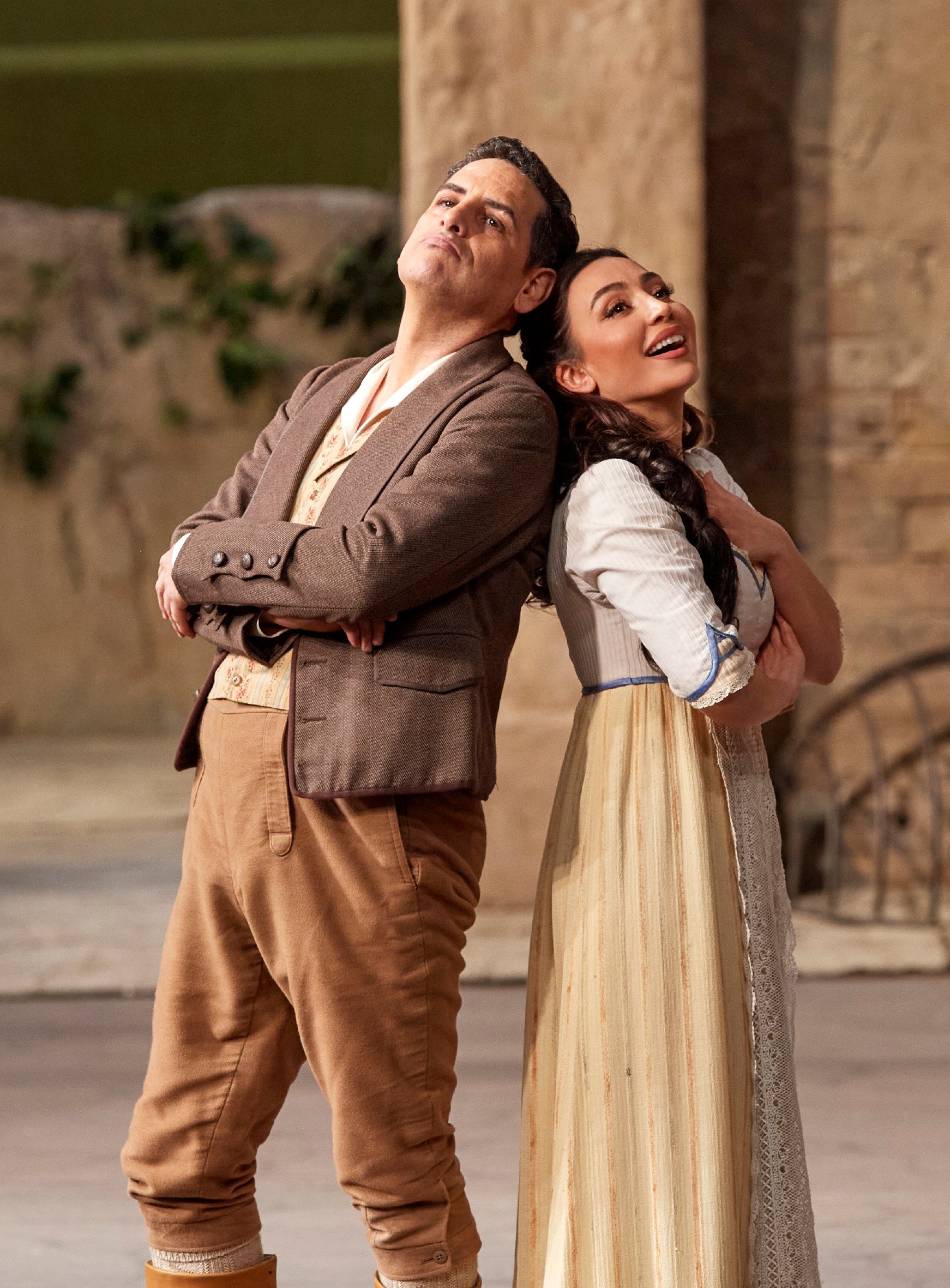 Ein Mann und eine Frau stehen Rücken an Rücken und lachen, in einer warmen, südländischen Bühnenkulisse.