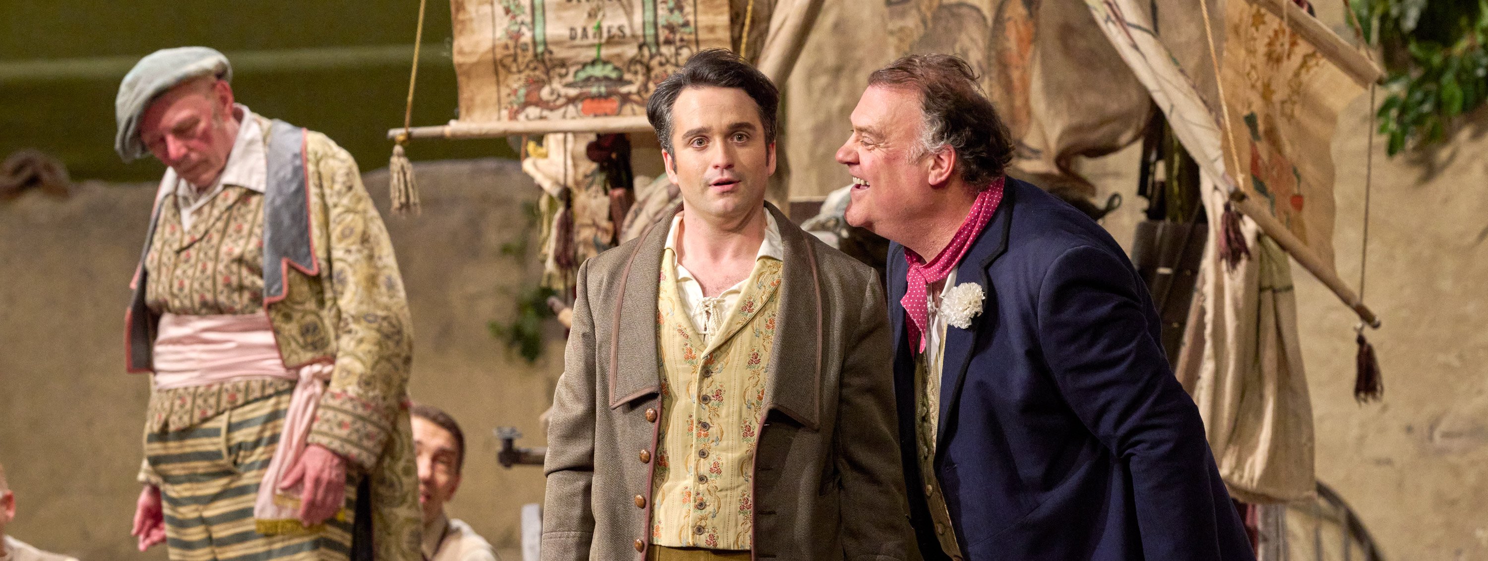 Ein Mann blickt überrascht, während ein älterer Herr ihm ins Ohr spricht, beide in historischer Kleidung vor einer Zeltkulisse.
