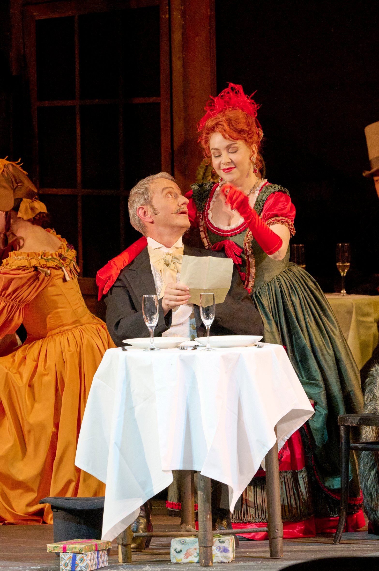 Eine elegant gekleidete Frau in Rot und Grün steht lächelnd an einem gedeckten Tisch und zeigt einem älteren Herrn ein Blatt Papier.