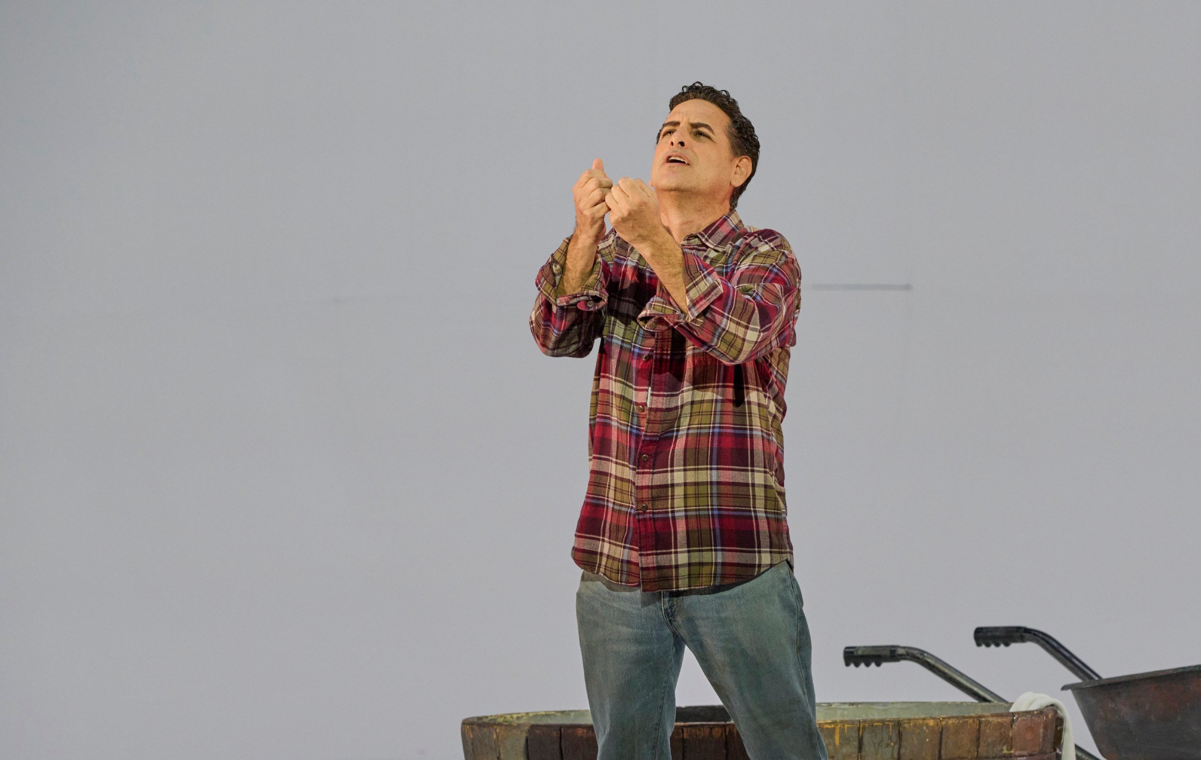 Ein Mann im karierten Hemd steht auf der Bühne und blickt mit erhobenen Händen eindringlich nach oben.