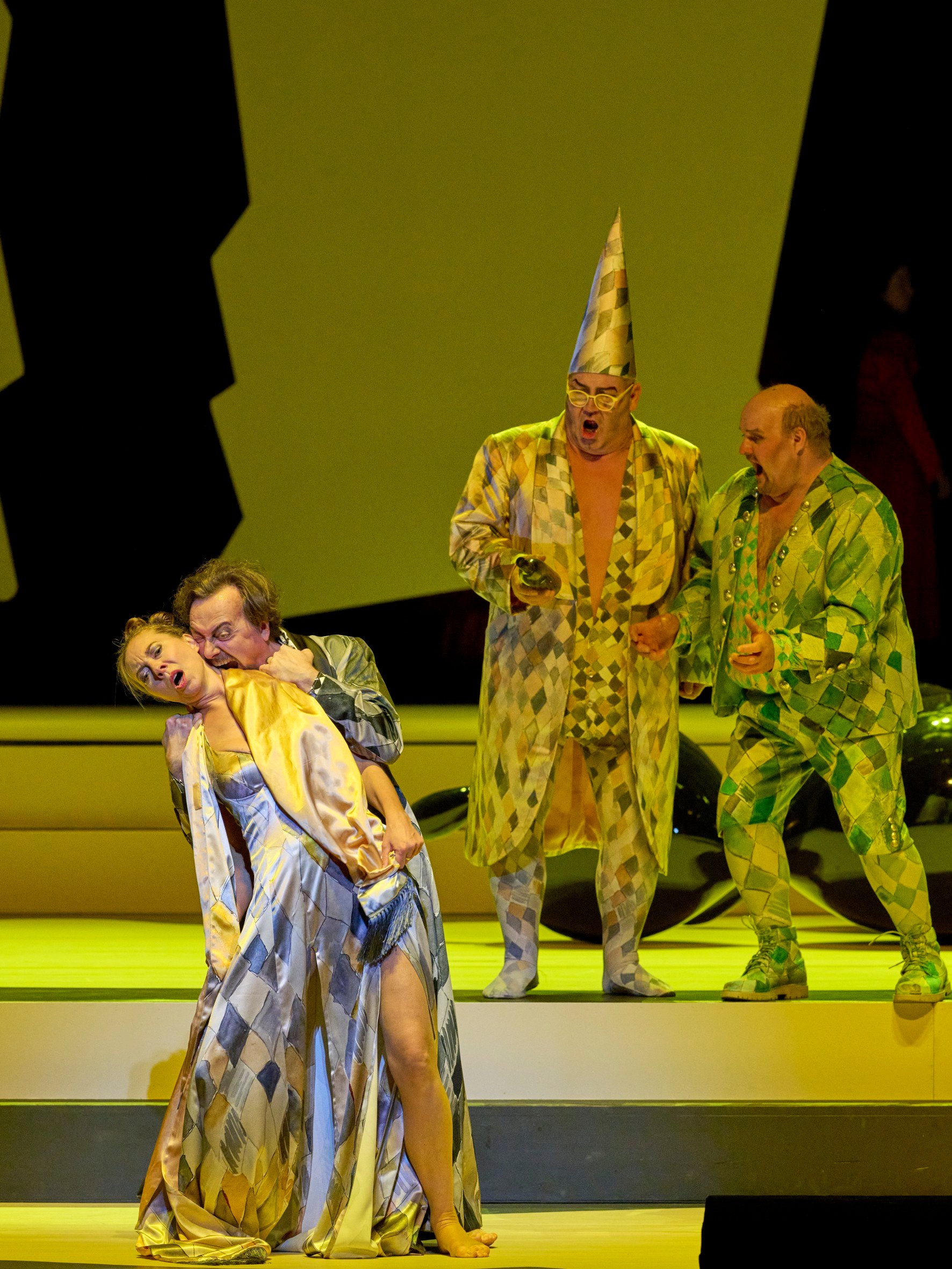 Ein Mann in buntem Anzug hält dramatisch eine Frau im Harlekin-Kleid, während zwei weitere Männer in auffälligen Kostümen im Hintergrund reagieren.