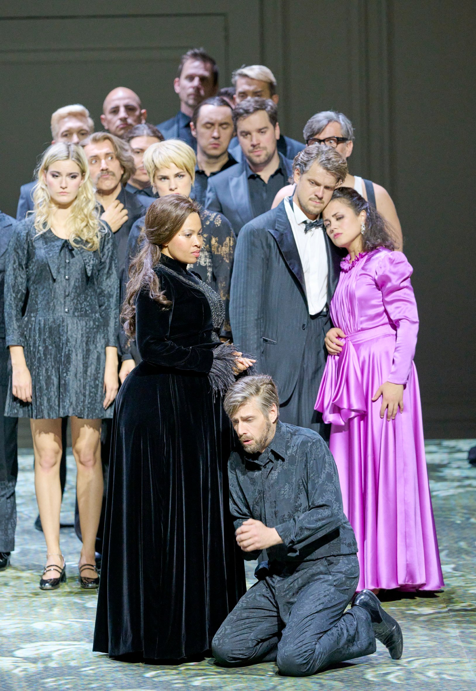 Eine Gruppe von Menschen steht dicht gedrängt beisammen; ein Mann im Anzug hält eine Frau im lila Kleid, die sich an seine Schulter lehnt.