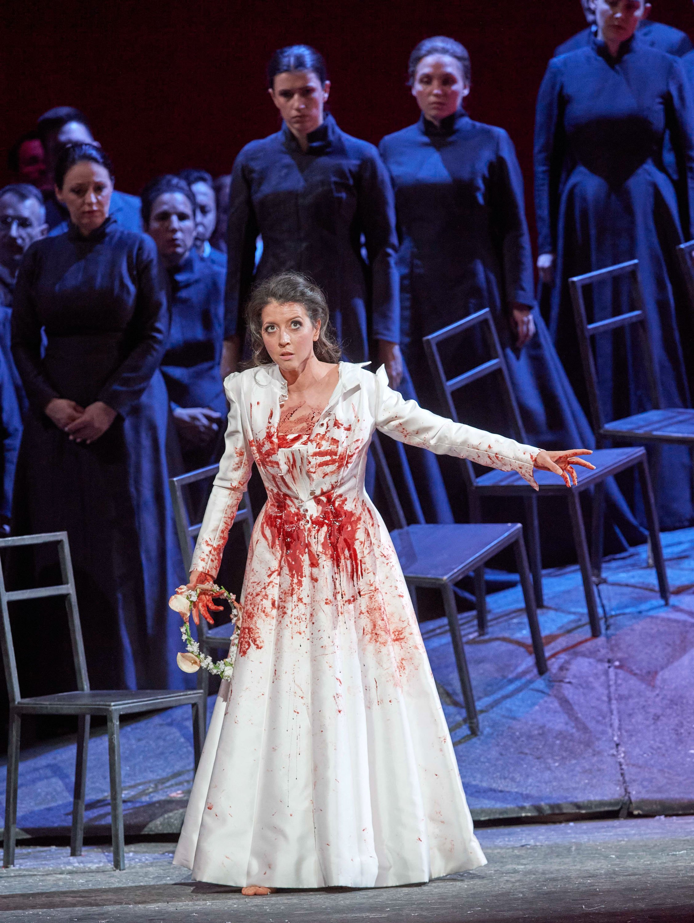 Eine Frau im blutbefleckten weißen Kleid steht im Zentrum einer dramatischen Opernszene, mit ausdrucksstarker Geste und intensiver Mimik.