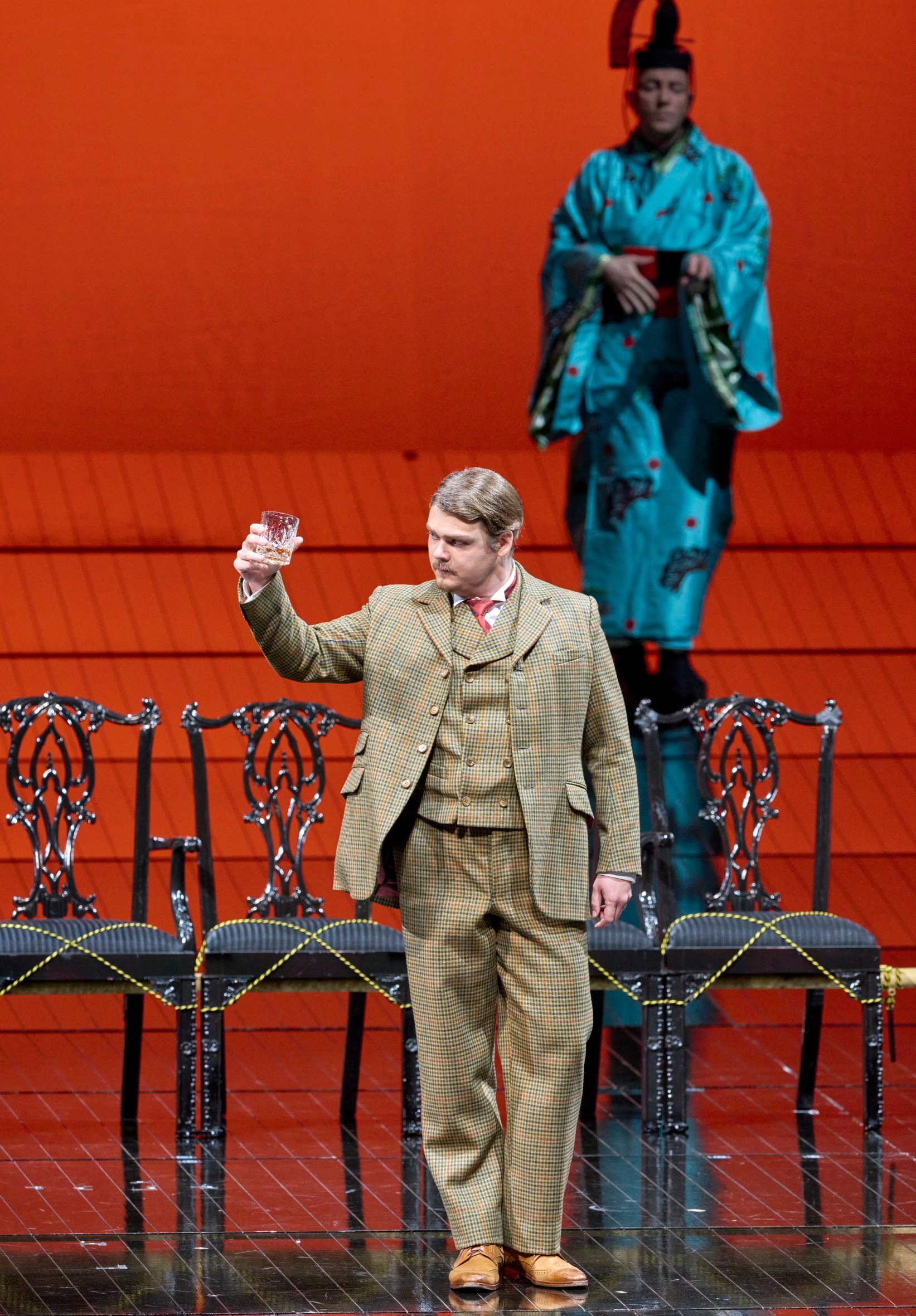 Ein Mann im Anzug hebt ein Glas auf einer roten Bühne, während im Hintergrund eine Frau im türkisfarbenen Kimono auf einer Treppe steht.