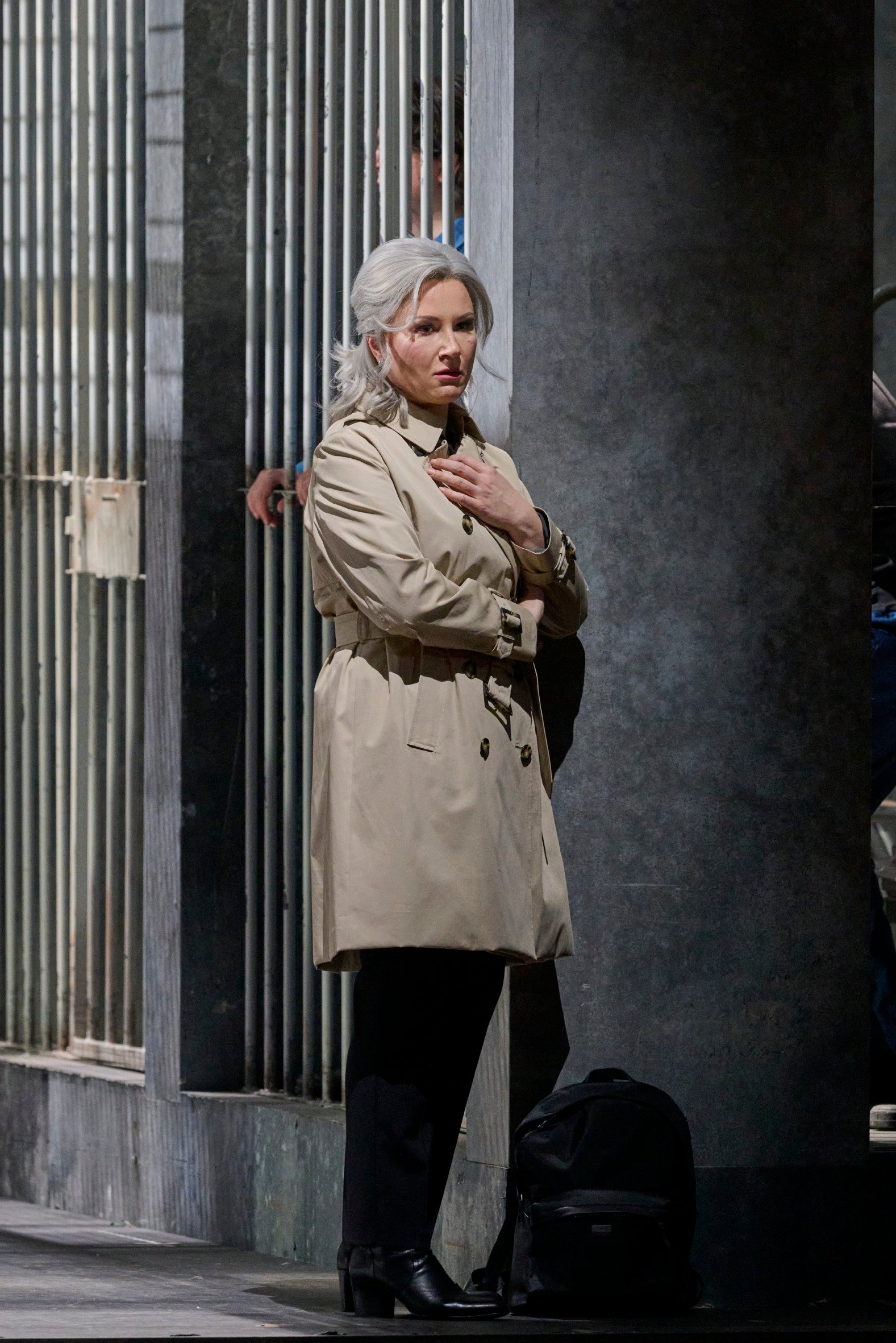 Eine Frau mit grauem Haar und beigem Trenchcoat steht nachdenklich an eine Wand gelehnt.