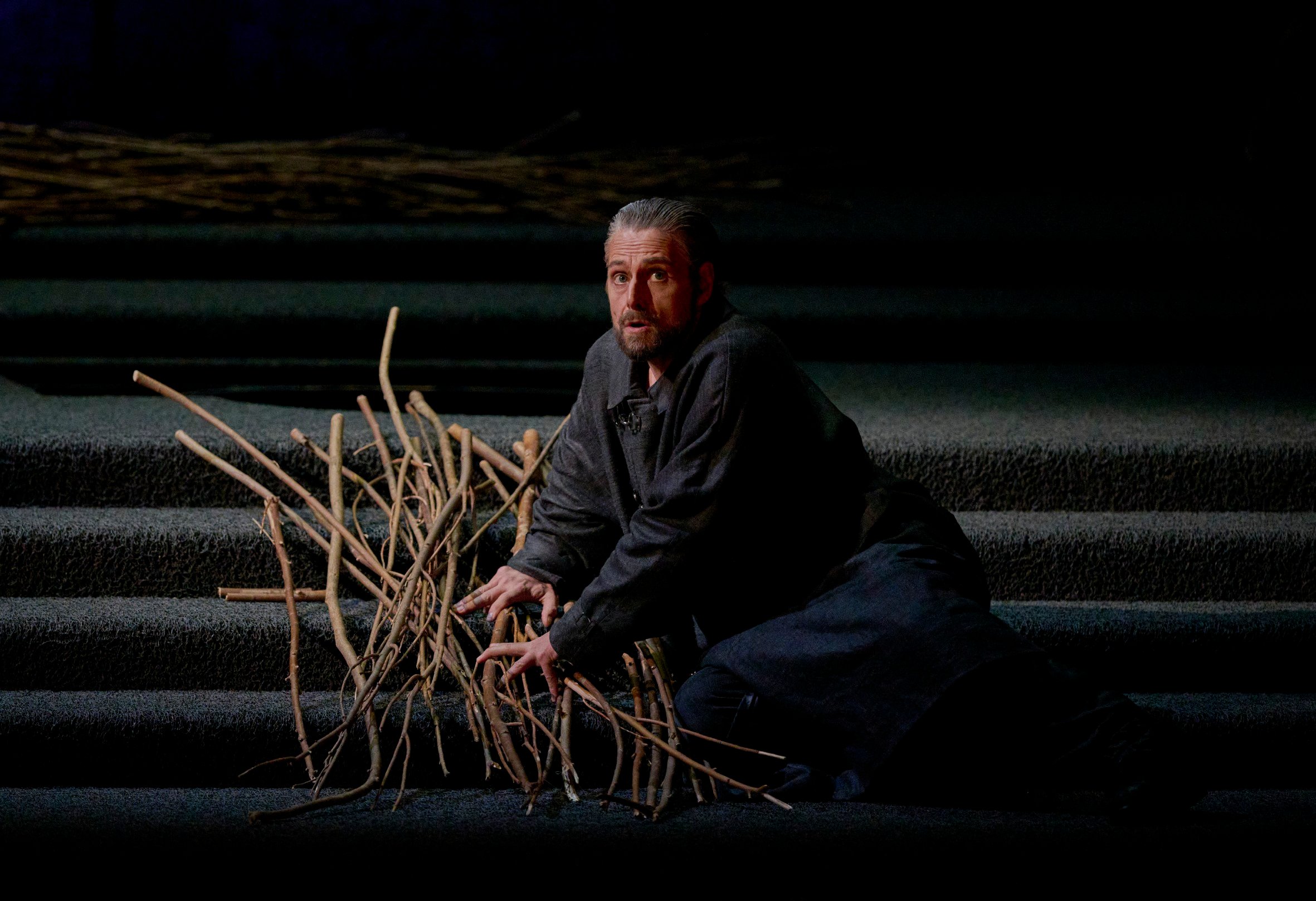 Ein Mann kniet auf einer dunklen Treppe und sammelt besorgt eine große Menge trockener Zweige.
