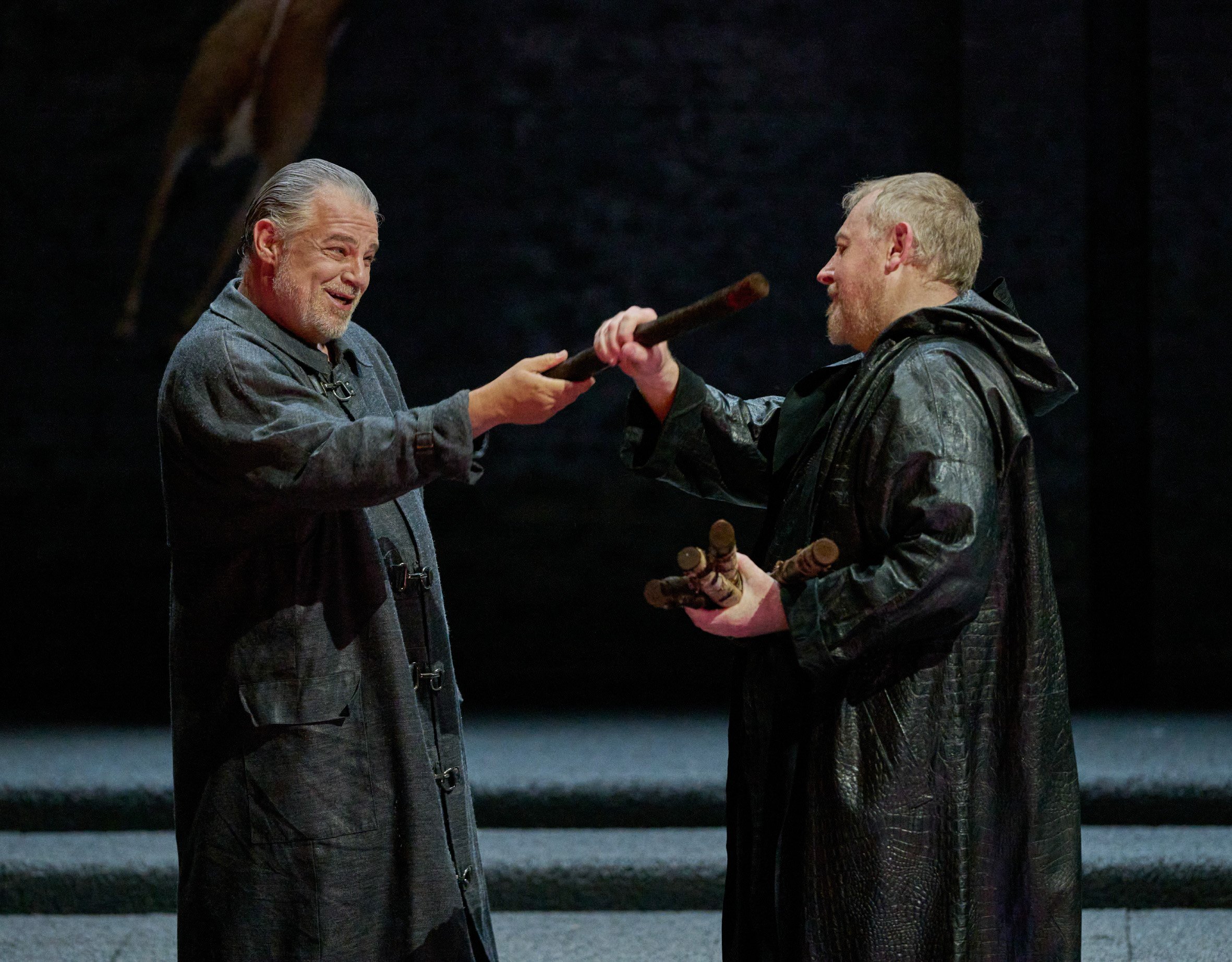 Zwei Männer in dunklem Gewand stehen sich gegenüber; der eine überreicht dem anderen einen Stab.