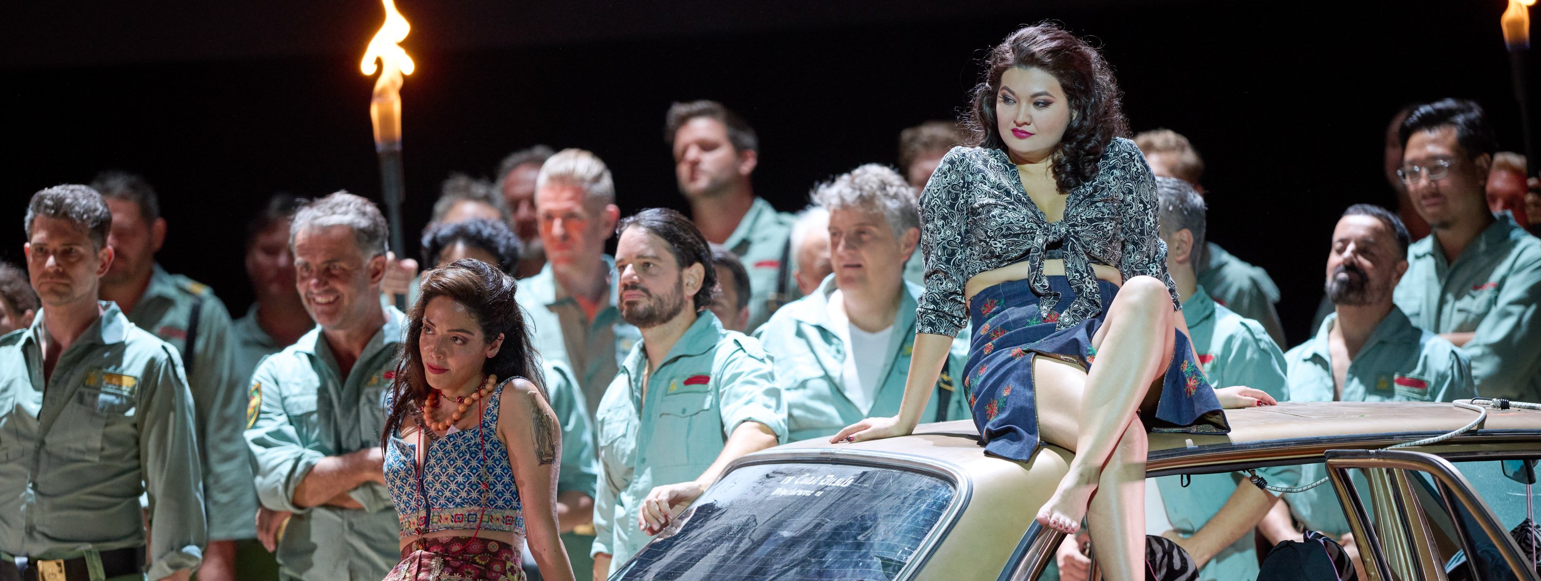 Eine Frau sitzt selbstbewusst auf einem Auto, während mehrere Männer in hellblauen Uniformen aufmerksam in eine Richtung blicken.