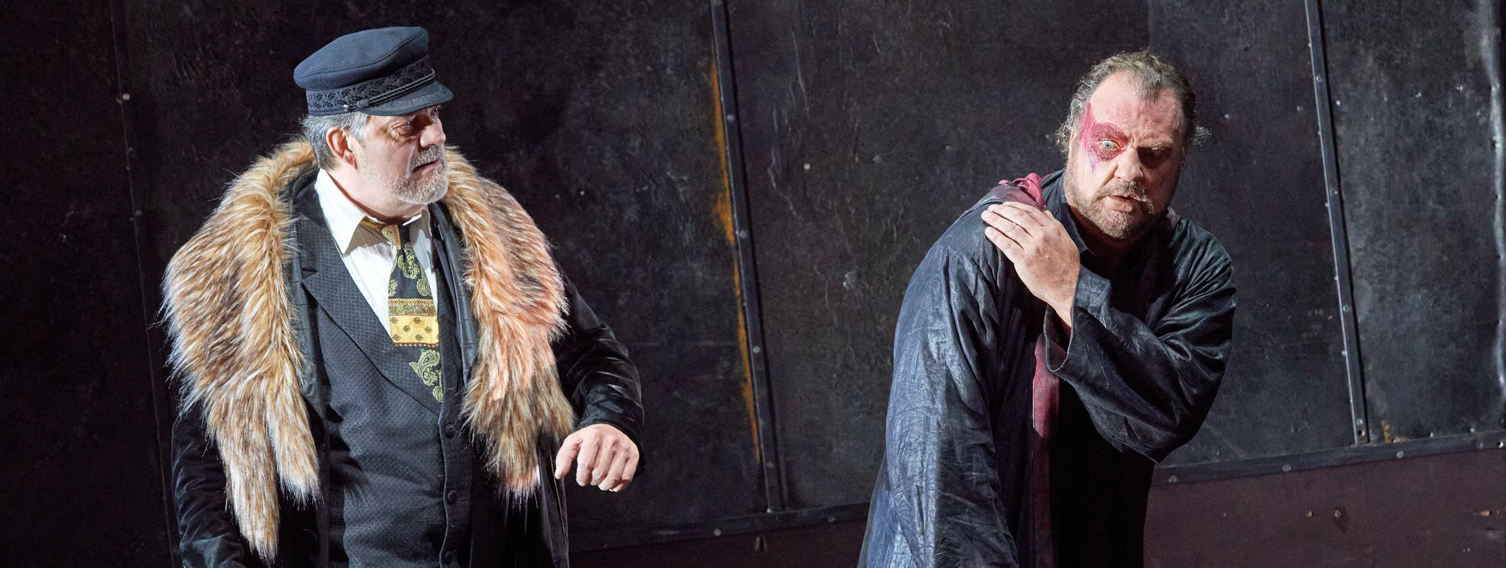 Zwei Männer in dunkler Kleidung stehen vor einer rostigen Metallwand; der eine trägt einen Pelzkragen, der andere wirkt verzweifelt.