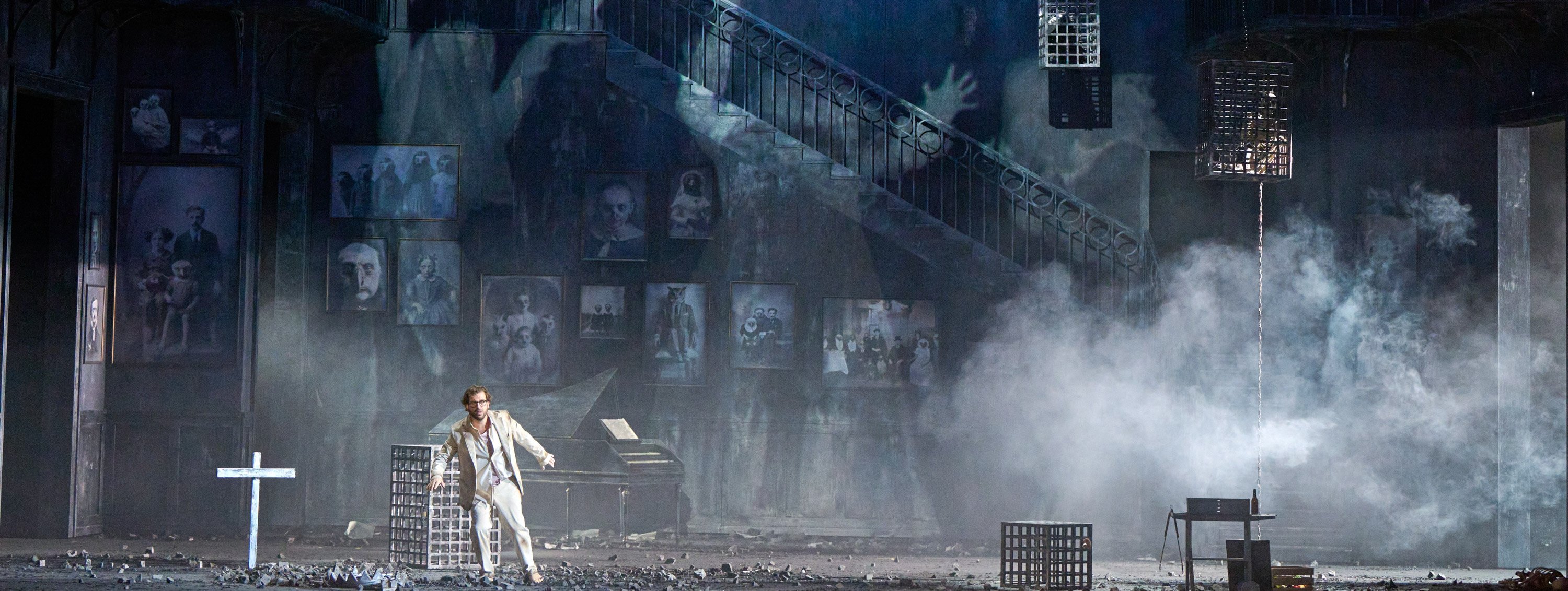 Eine geisterhafte Bühnenkulisse mit rauchigem Nebel, düsteren Porträts und riesigen, transparenten Erscheinungen über einer Treppe; eine weiß gekleidete Figur steht links.