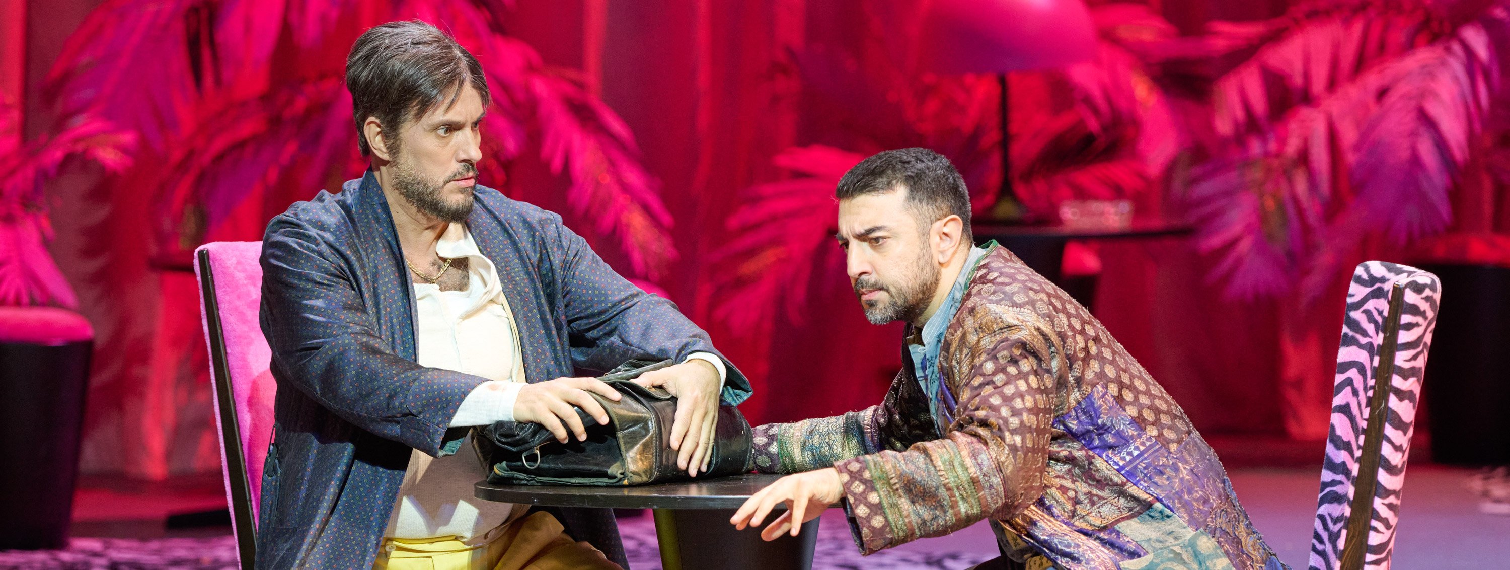 Zwei Männer in gemusterten Gewändern sitzen sich an einem Tisch gegenüber und streiten angespannt um eine Tasche, vor einem pink beleuchteten Hintergrund mit Palmen.