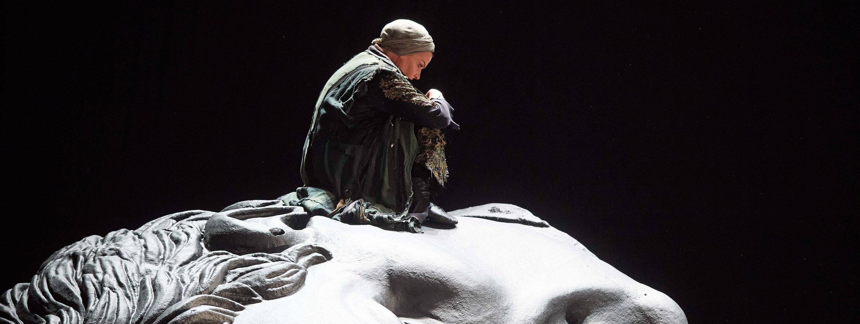 Eine in dunkle Stoffe gehüllte Person sitzt nachdenklich auf einer weißen, skulpturalen Oberfläche in Form eines Gesichts.