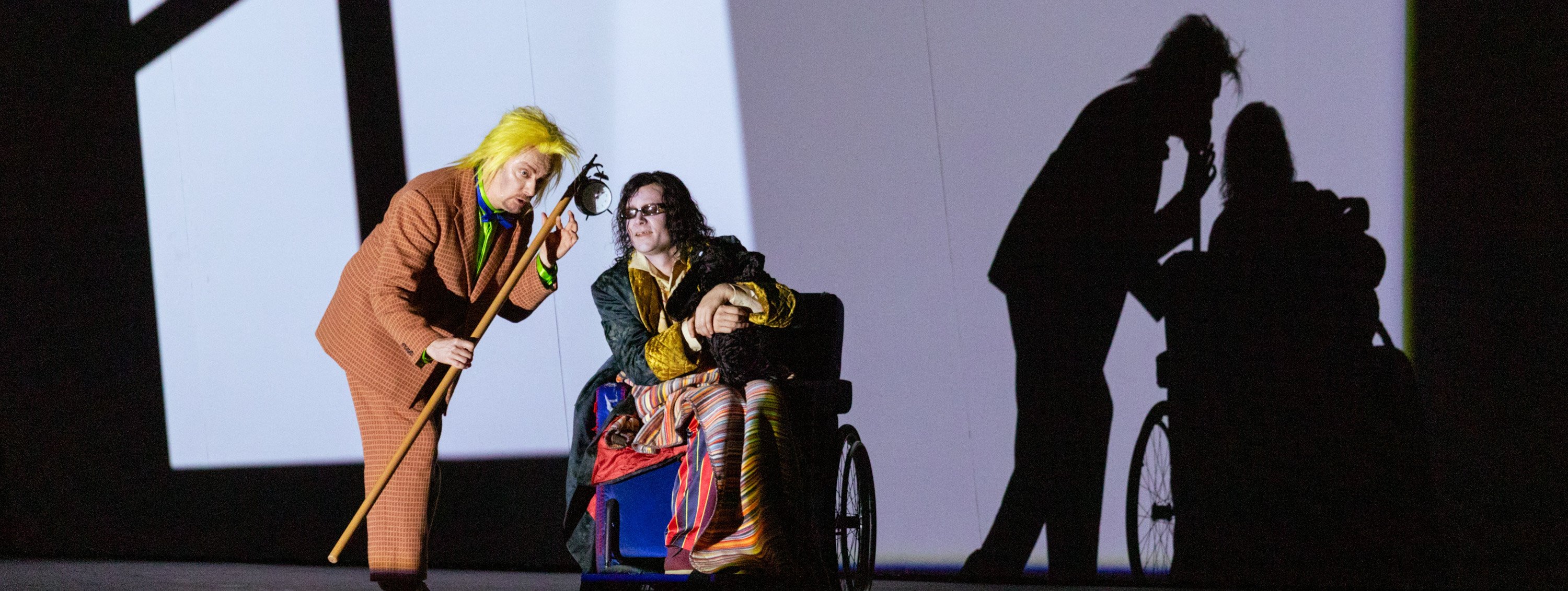 Eine Person mit knallgelber Perücke spielt Laute neben einer in bunte Stoffe gehüllten Figur im Rollstuhl, während eine große Schattenprojektion die Szene verdoppelt.