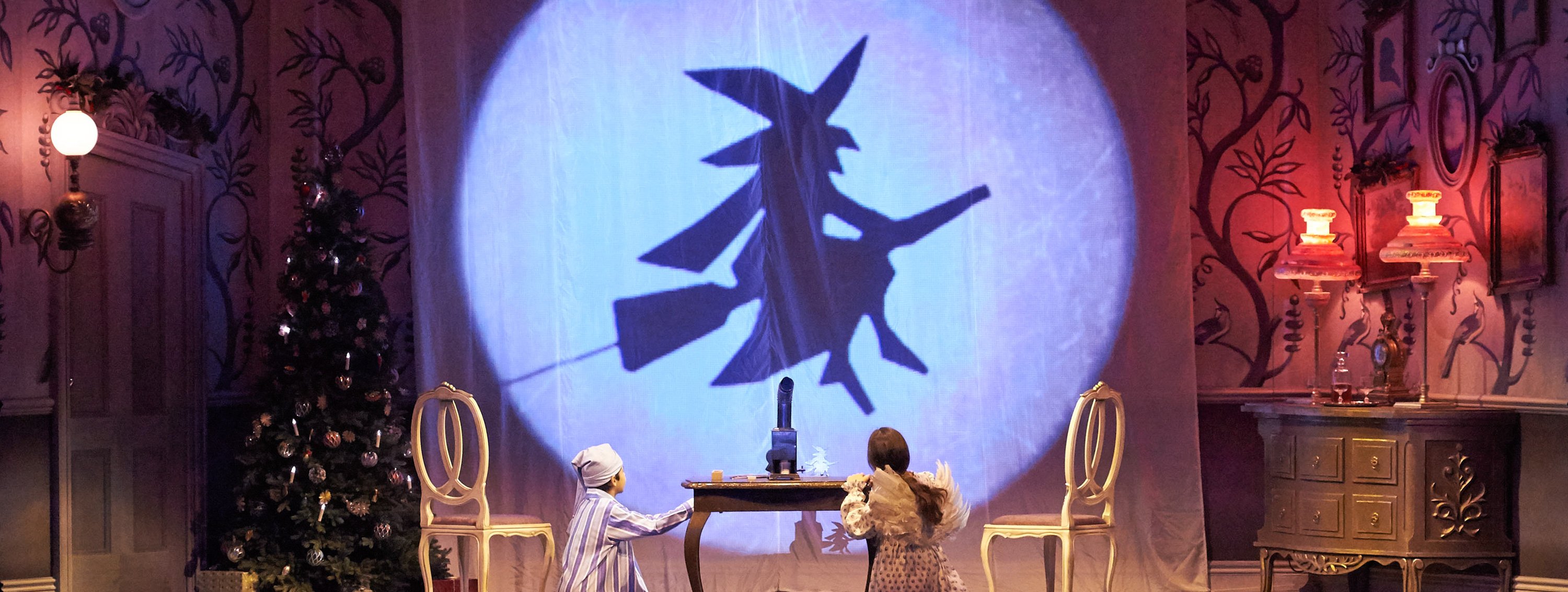 Zwei Kinder in Kostümen blicken auf eine Wandprojektion, die den Schattenschnitt einer fliegenden Hexe auf einem Besen zeigt.