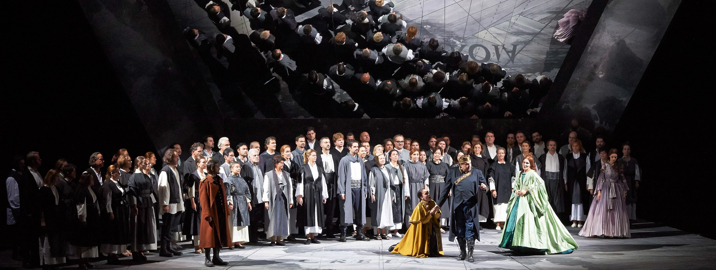 Ein großer Chor in einheitlich grauen Kostümen steht auf der Bühne, während zwei Solisten im Vordergrund agieren und eine Spiegeldecke die Szene verdoppelt.