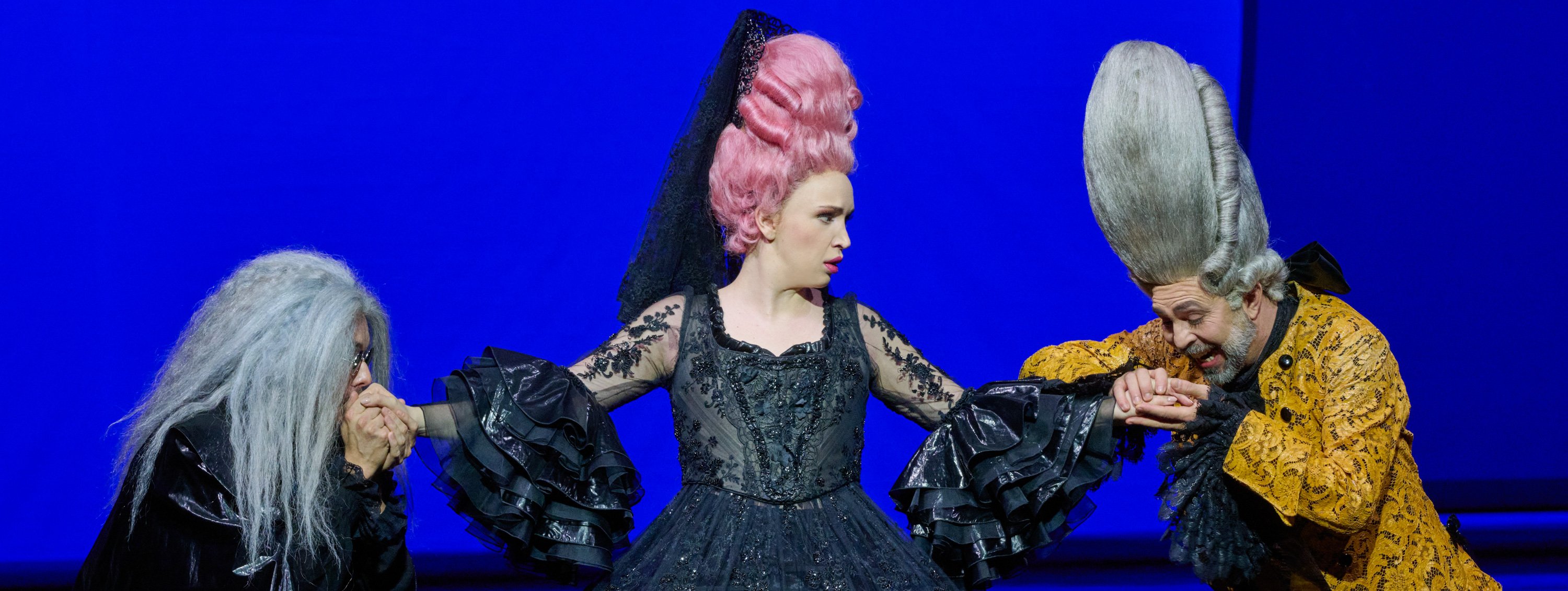 Eine Frau mit hochgesteckter rosa Perücke und schwarzer Spitze trägt ein ausladendes schwarzes Kleid vor tiefblauem Hintergrund und blickt energisch zur Seite.