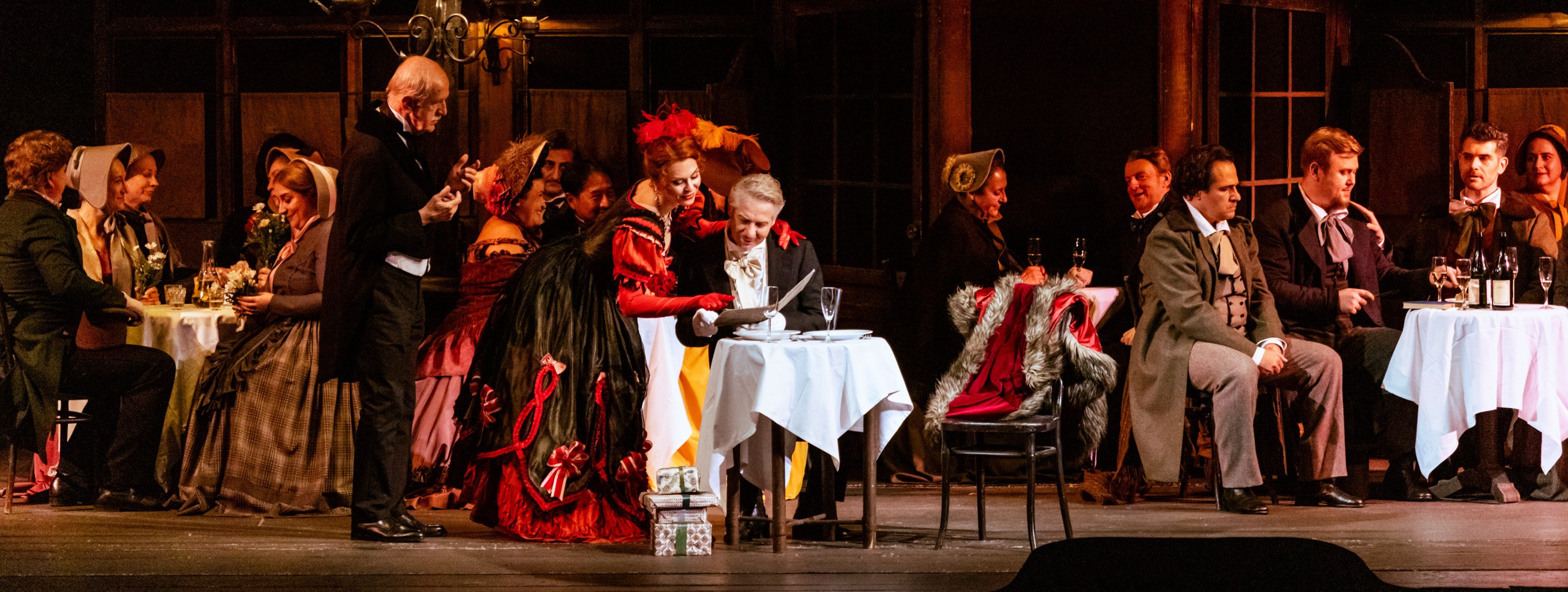 Ein elegant gedeckter Tisch auf der Bühne wird von einem festlich gekleideten Mann betrachtet, während eine Frau in rotem Kleid ihn charmant umgarnt und andere Figuren in historischen Kostümen zusehen.