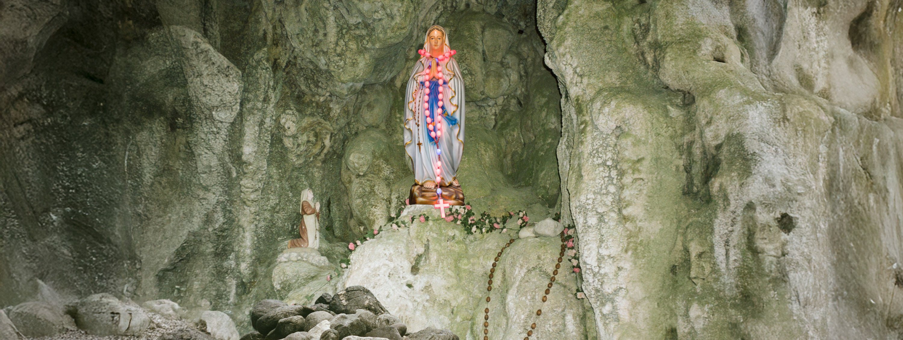 Eine bunt geschmückte Marienstatue mit Rosenkränzen steht in einer Felsgrotte, daneben eine kleinere kniende Figur.