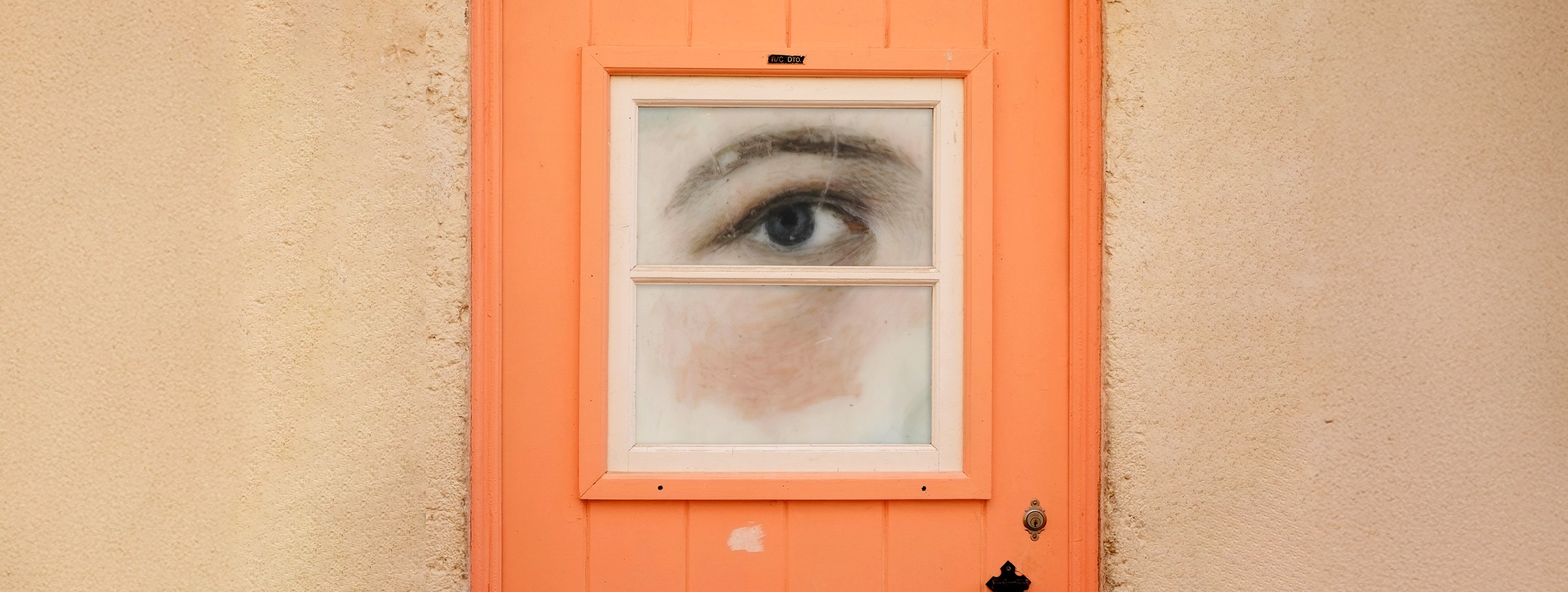 Ein gemaltes menschliches Auge blickt durch das kleine Fenster einer orangefarbenen Tür.