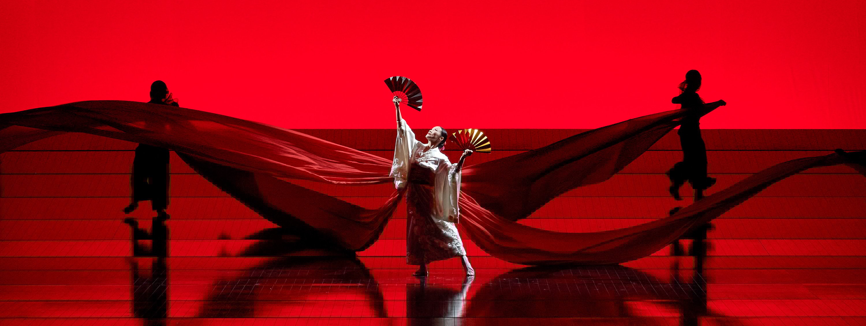 Vor einem leuchtend roten Hintergrund tanzt eine Person in weißem Gewand mit zwei Fächern, während sich ein roter Stoff über die Bühne spannt.