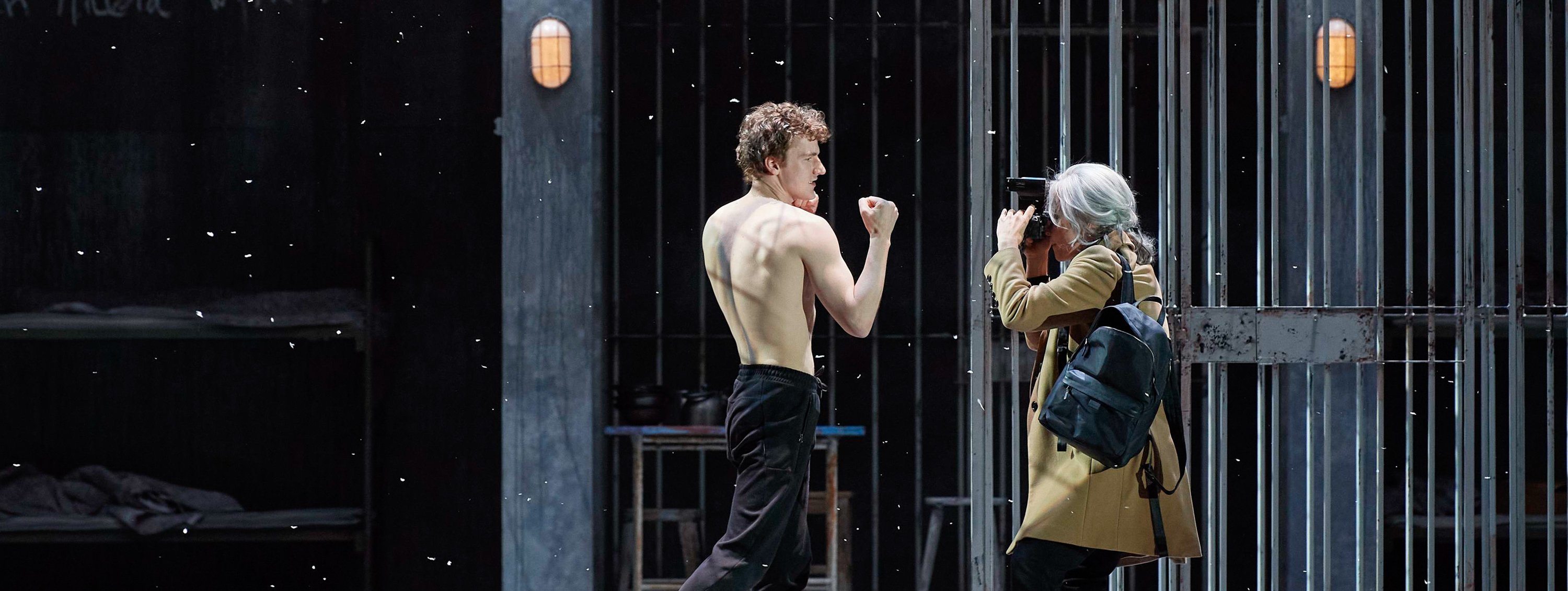 Ein junger Mann mit nacktem Oberkörper posiert mit geballter Faust in einem dramatischen Moment, während ein älterer Fotograf in einem beigen Mantel ein Foto von ihm macht. Im Hintergrund ist ein dunkler, gitterartiger Bühnenaufbau zu sehen.