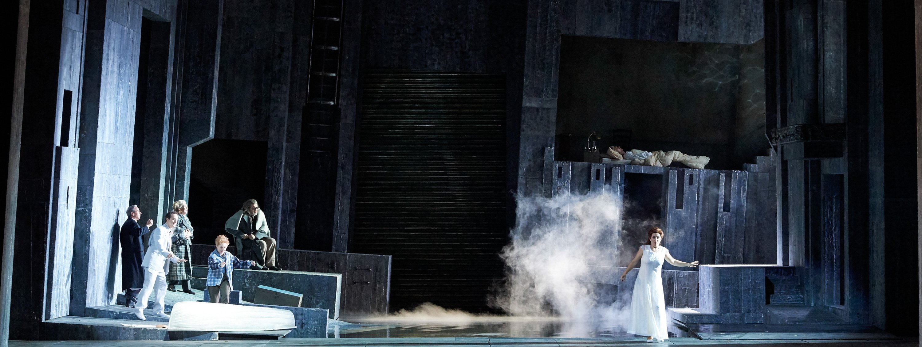 Eine weiß gekleidete Frau tritt aus dem Nebel auf eine düstere Bühne, während rechts oben eine Gestalt in einem Bett liegt und links eine Gruppe Menschen sie beobachtet.