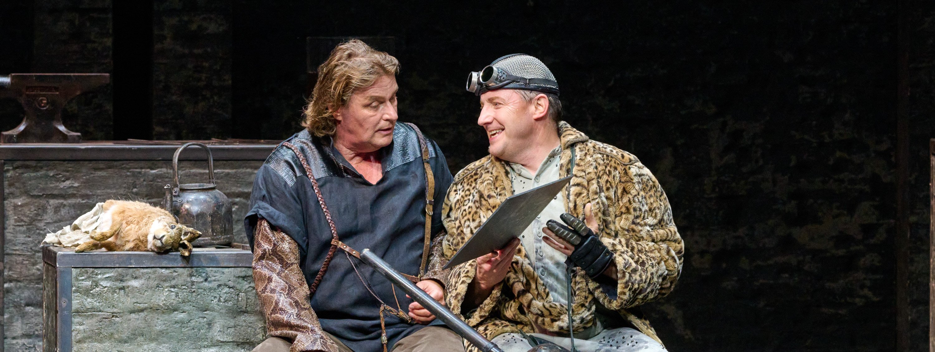 Zwei Männer sitzen nebeneinander, während der eine dem anderen mit einem fröhlichen Gesichtsausdruck ein Blatt Papier zeigt.