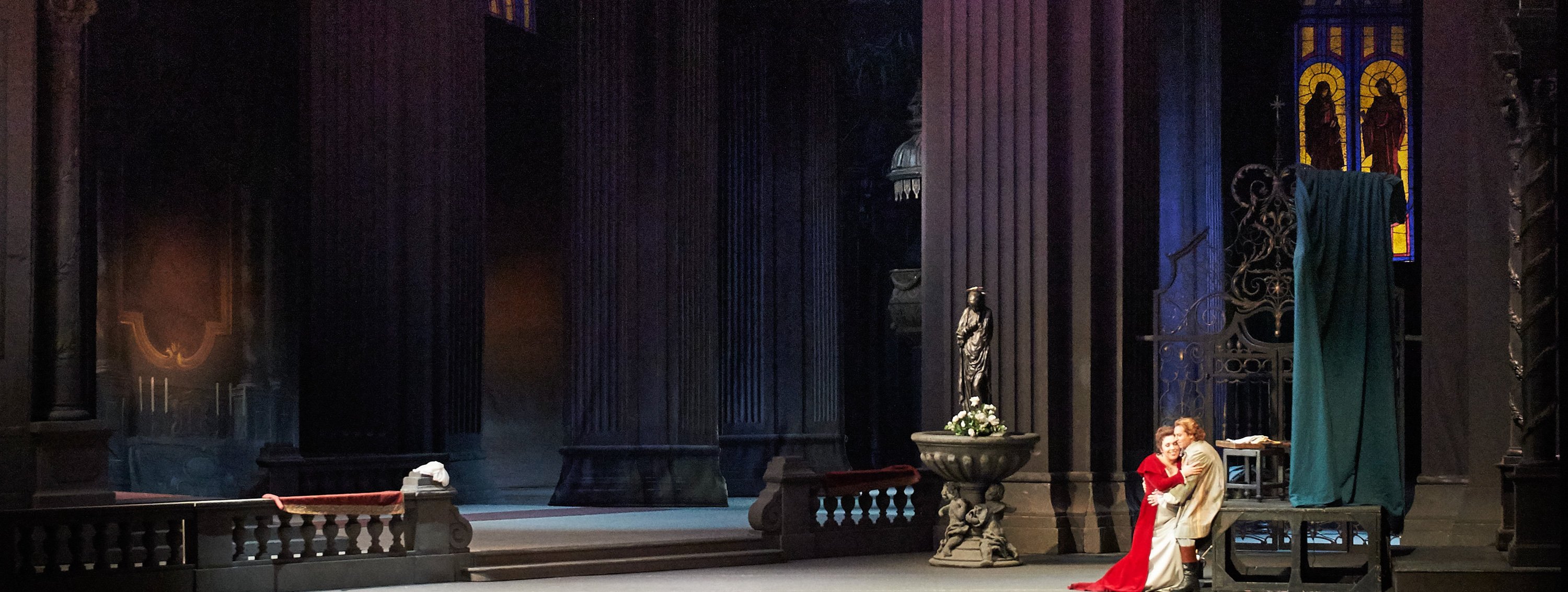 Ein leidenschaftliches Liebespaar steht in inniger Umarmung im rechten Vordergrund einer monumentalen, düsteren Kirchenkulisse, während farbiges Licht durch das hohe Buntglasfenster fällt.