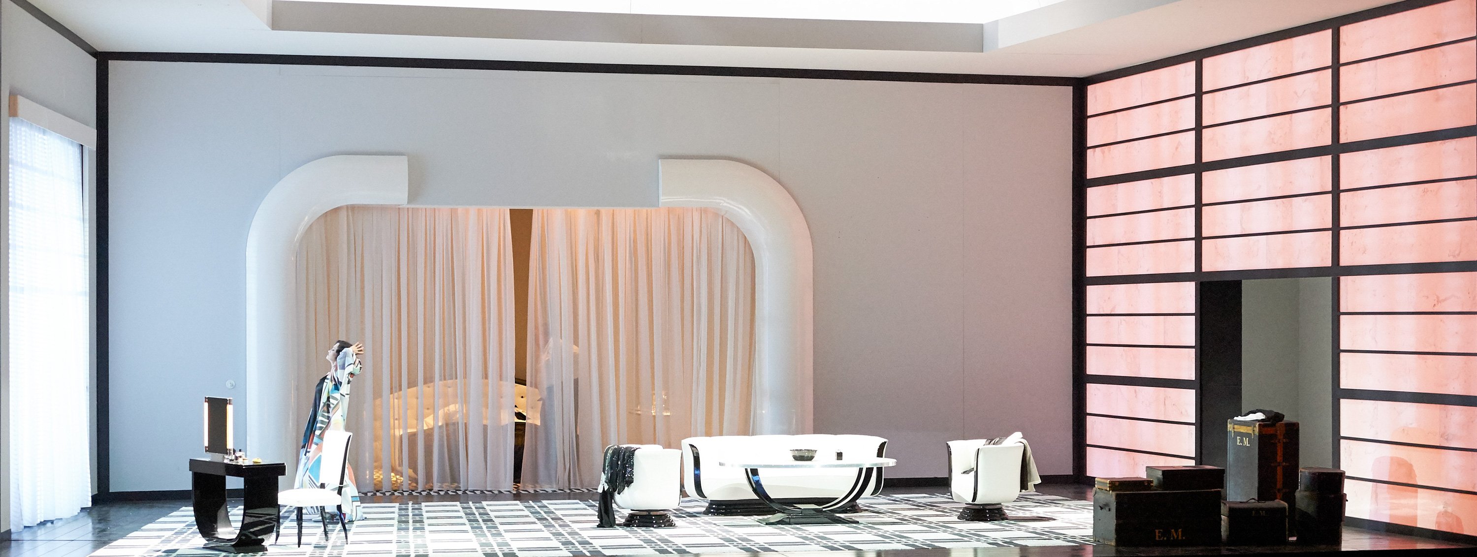 Eine elegant eingerichtete Bühne zeigt ein modernes Zimmer mit weißen Möbeln, rosa Wandelementen und einer Person im farbenfrohen Gewand, die sich streckt.