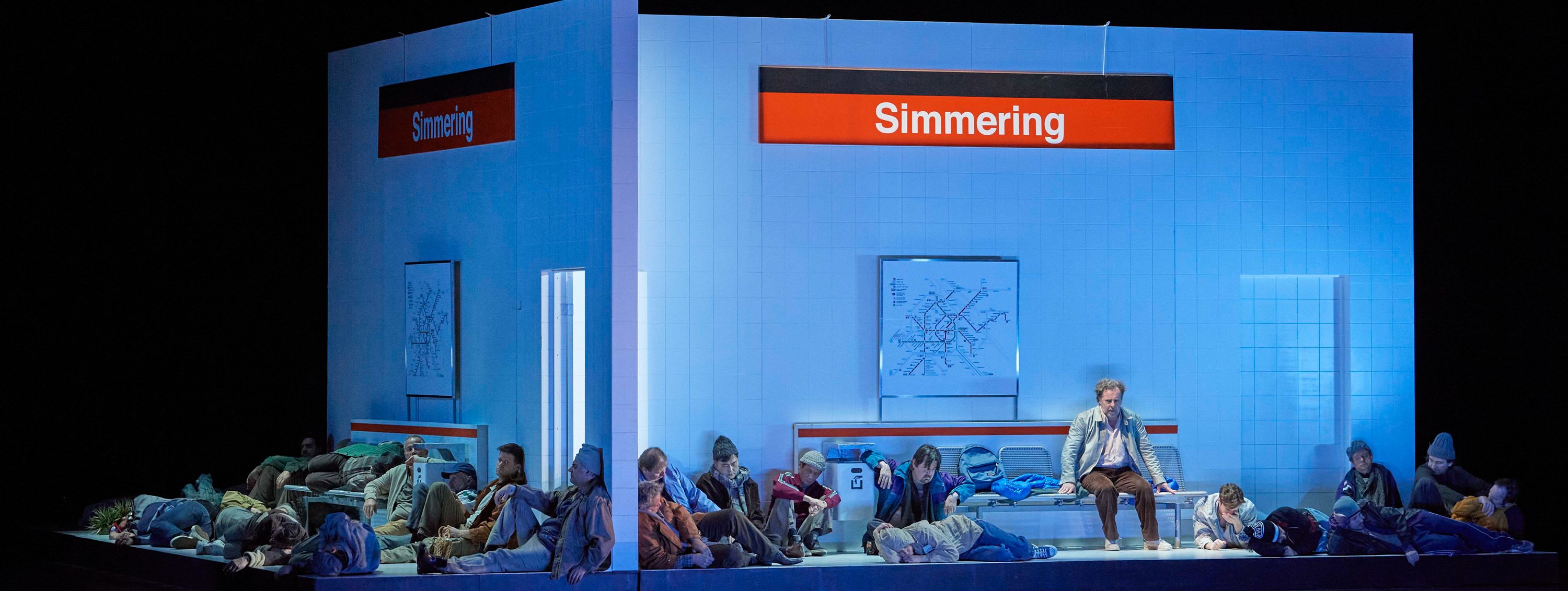 Eine Gruppe von Personen sitzt auf dem Boden in einer U-Bahn-Station mit dem Schild "Simmering" im Hintergrund.