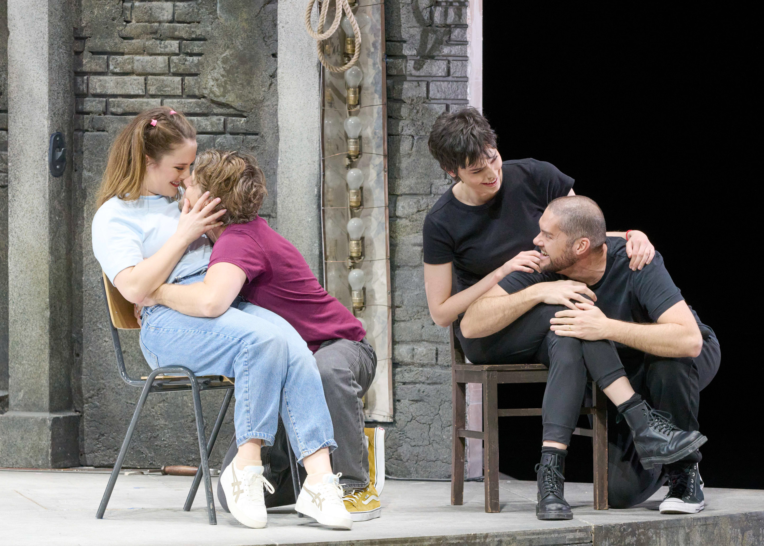 Zwei Paare sitzen in entspannter Atmosphäre auf Stühlen vor einer Backsteinwand; links umarmen sich zwei Personen liebevoll, rechts wird ein intimes Gespräch geführt.