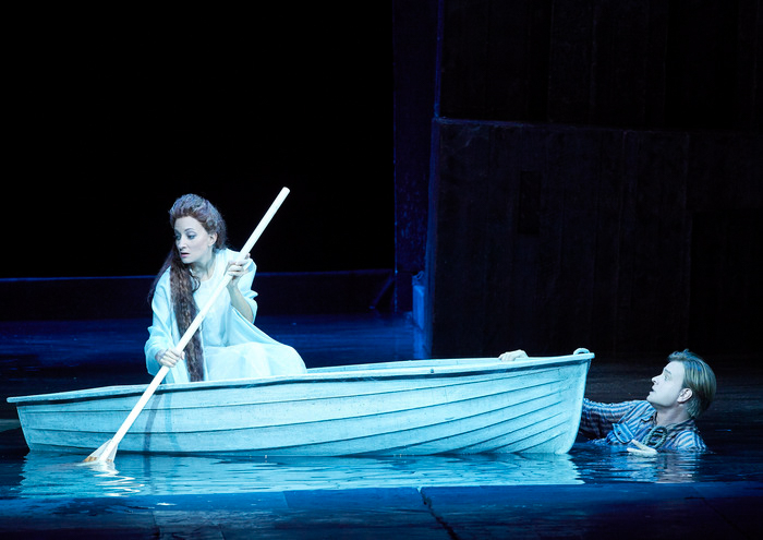 Eine Frau mit langem Haar und weißem Kleid rudert allein in einem kleinen Boot durch eine blaue Bühnenlandschaft.