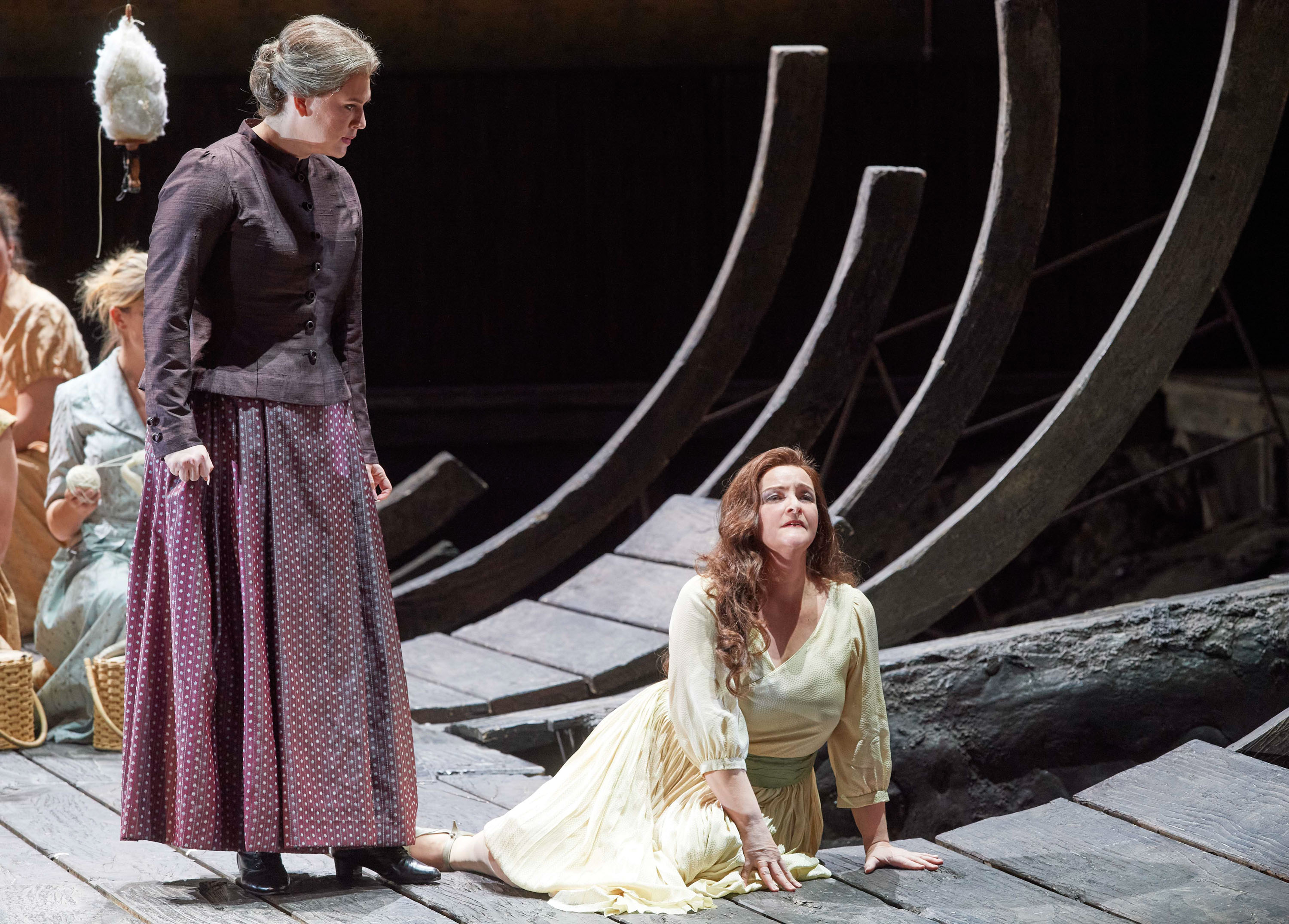 Eine ältere Frau blickt streng auf eine jüngere Frau in hellem Kleid, die verzweifelt auf dem Boden kniet, während andere im Hintergrund sitzen.