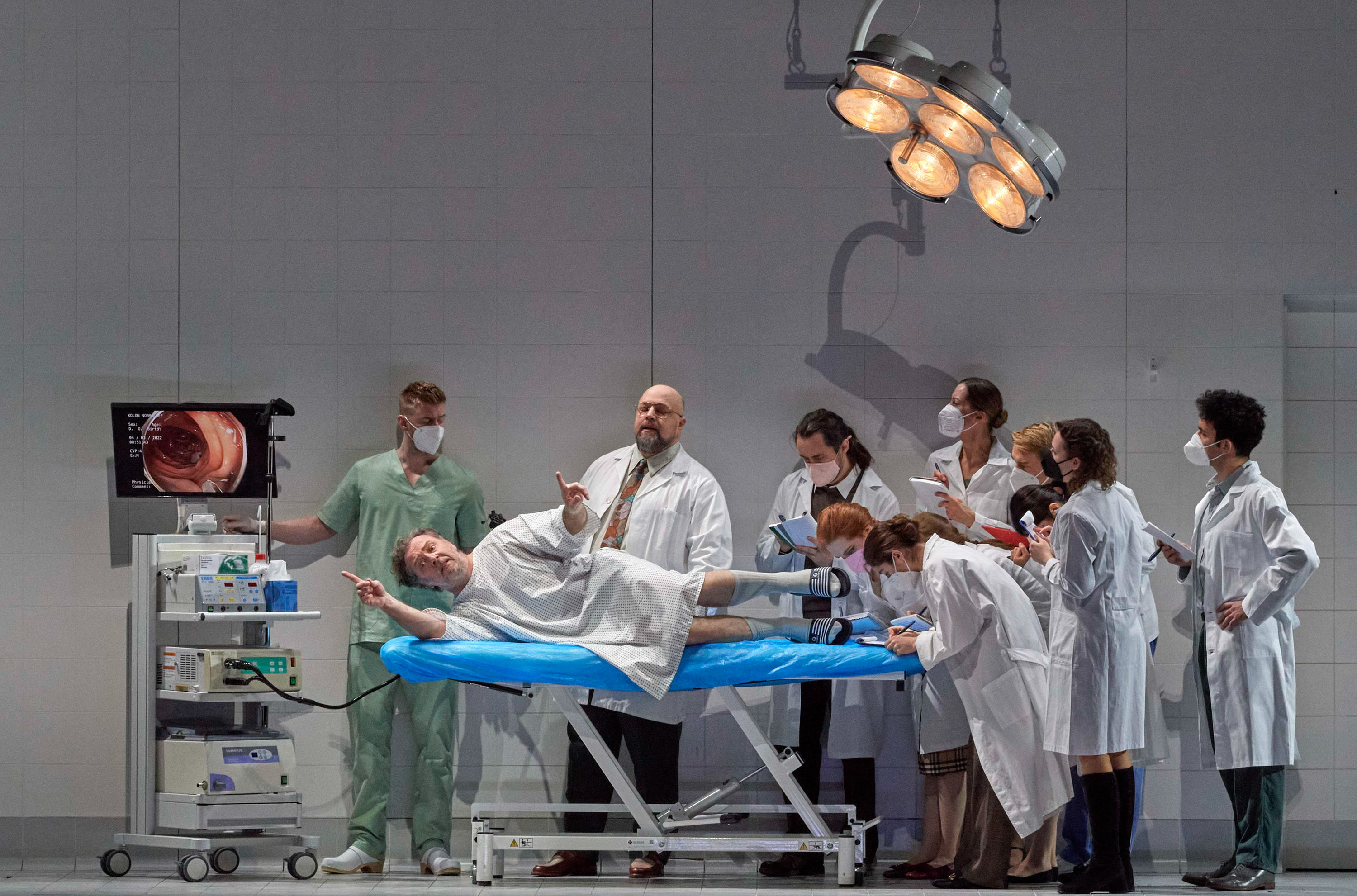 In einem Behandlungszimmer stehen verschiedene Personen, eine davon liegt auf einer Liege im Patientenkittel und blickt nach vorne.