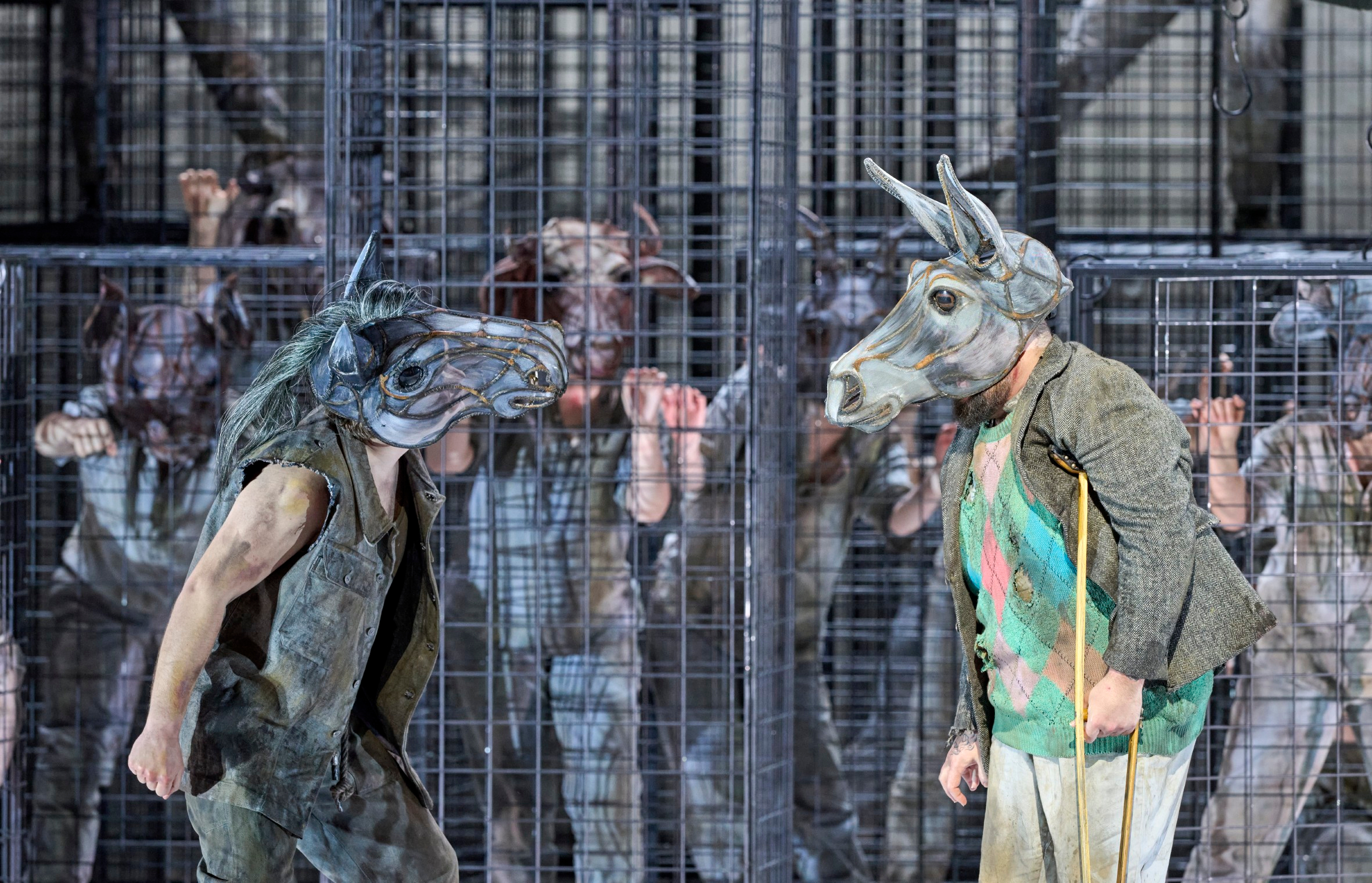 Eine Person mit einer Pferdemaske bewegt sich dynamisch vor einem Käfig, in dem mehrere Figuren mit Tiermasken gefangen sind.