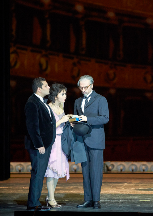 Zwei Männer im Anzug und eine Frau im hellen Kleid stehen auf einer Bühne; ein Mann hält ihre Hände.