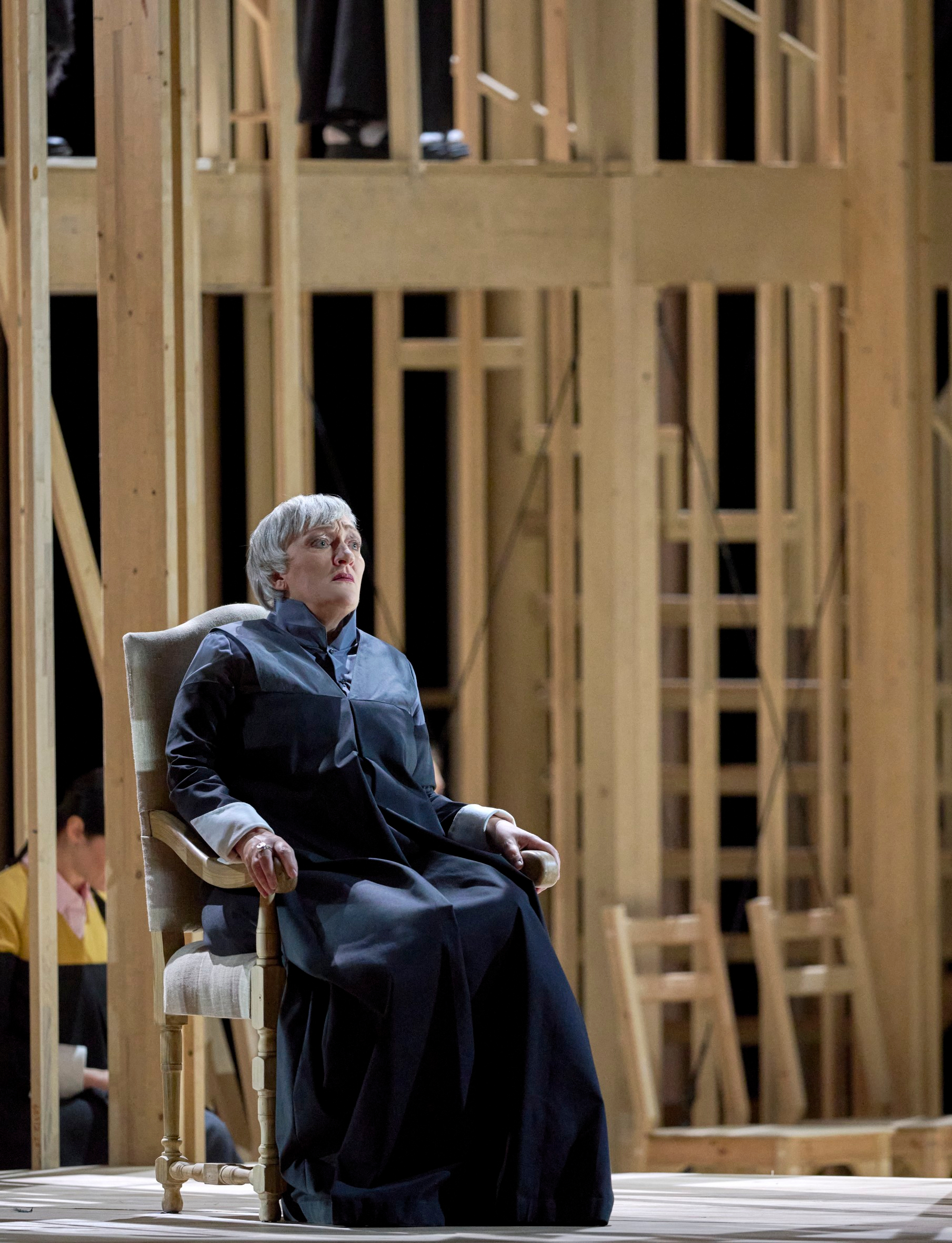 Eine ältere Frau in dunkler Kleidung sitzt nachdenklich auf einem Sessel vor einem Bühnenbild aus hellem Holzgerüst.