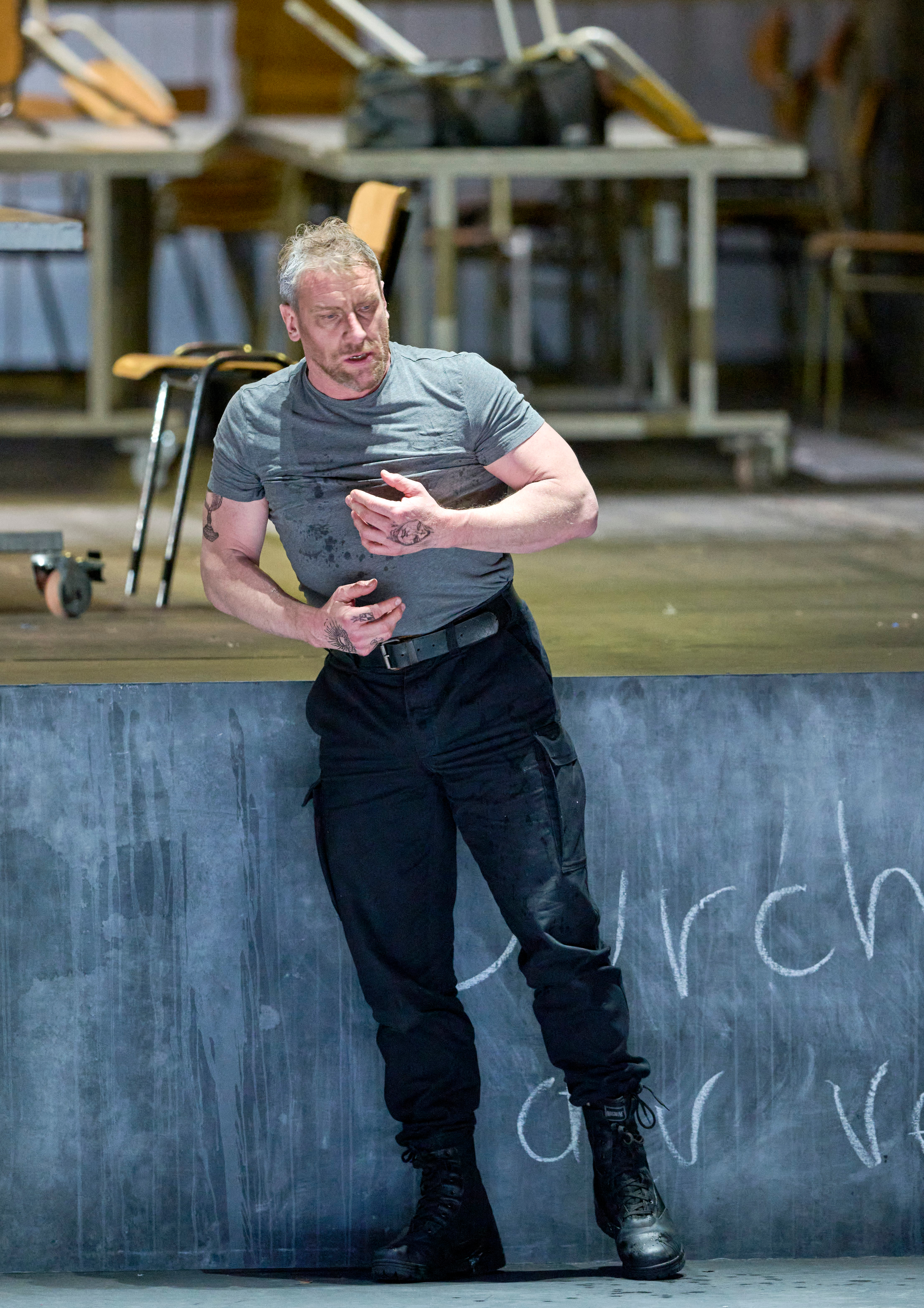 Ein verletzter Mann in grauem T-Shirt lehnt sich mit blutverschmierter Hand an eine Wand, im Hintergrund stehen umgestürzte Stühle.