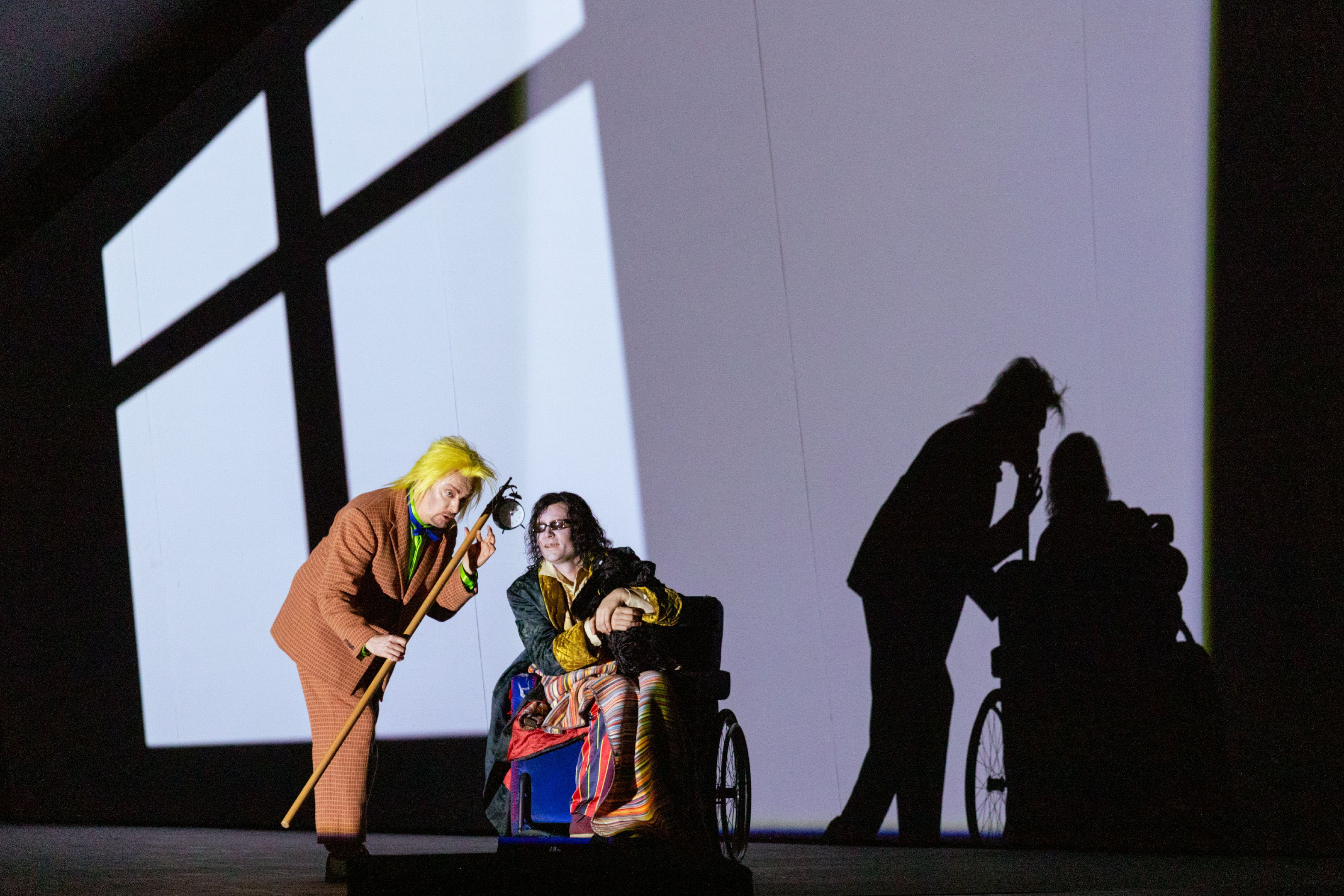 Eine Figur mit gelber Perücke beugt sich zu einer Person im Rollstuhl, beide vor einer Wand mit markanten Schattenprojektionen.