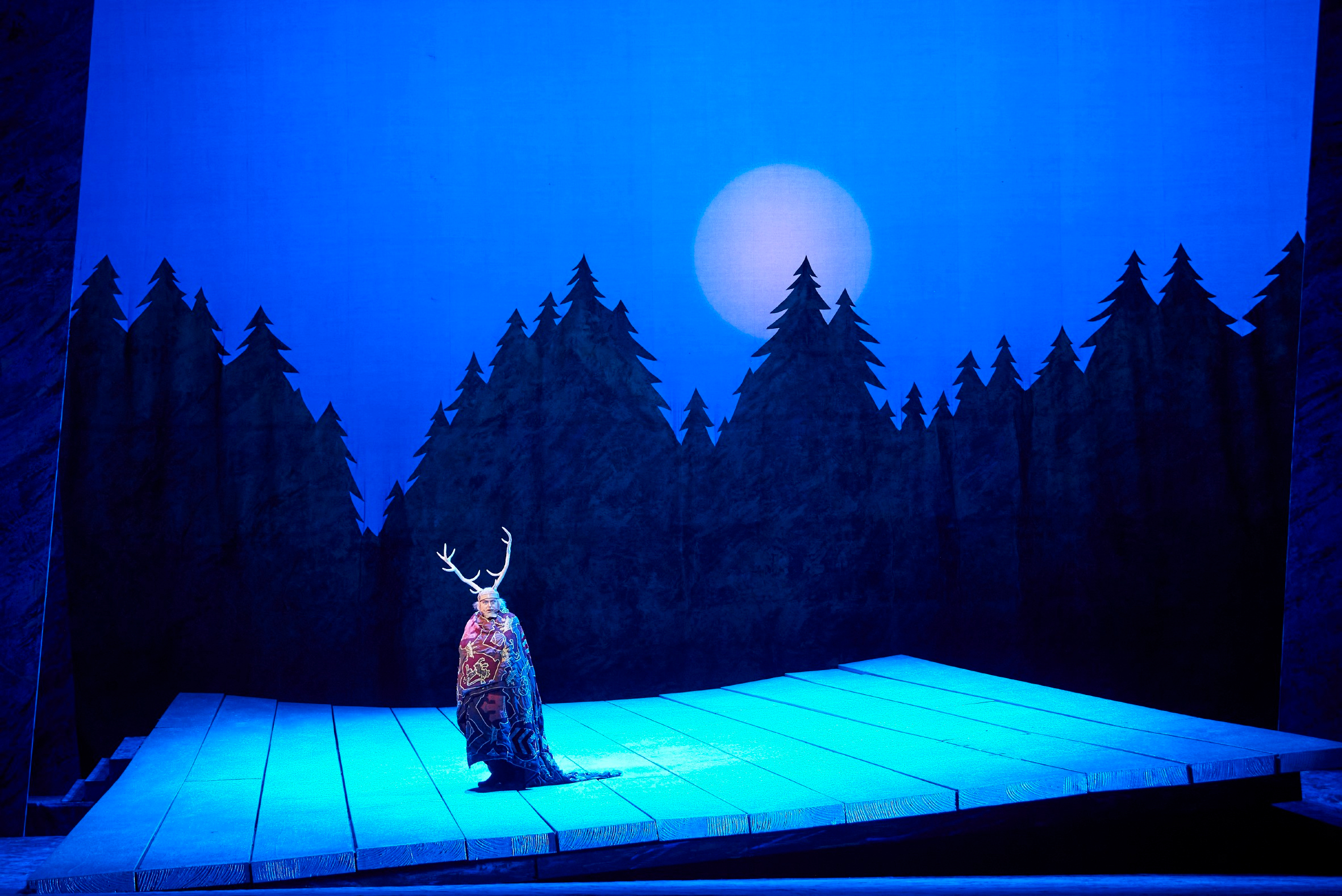 Eine Figur mit Hirschgeweih steht allein auf einer in Blau beleuchteten Bühne vor einer Kulisse mit Tannenwald und Vollmond.