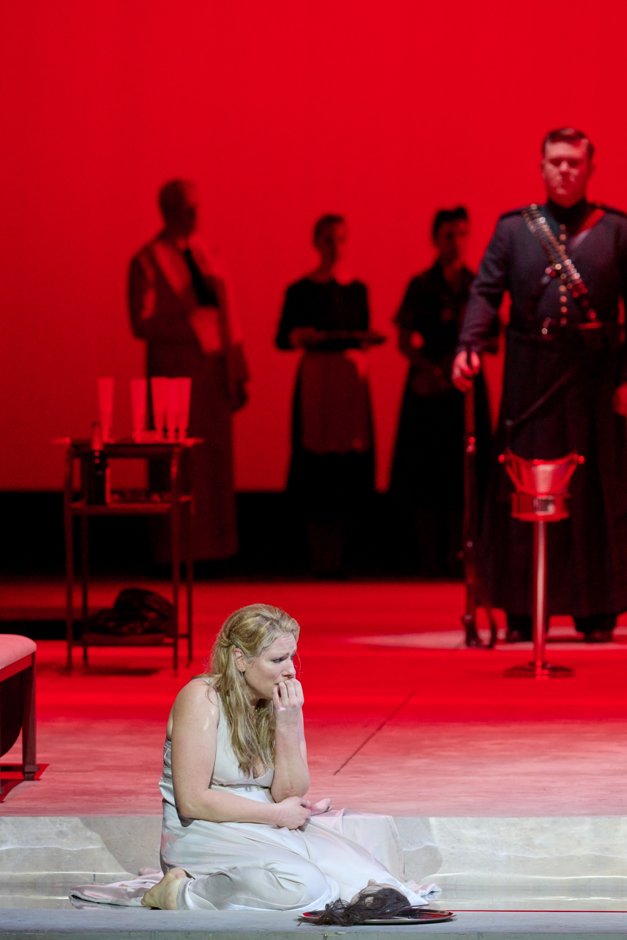 Eine Frau im weißen Kleid sitzt verzweifelt auf dem Boden einer rot ausgeleuchteten Bühne, umgeben von medizinischem Besteck und im Hintergrund stehenden Personen.