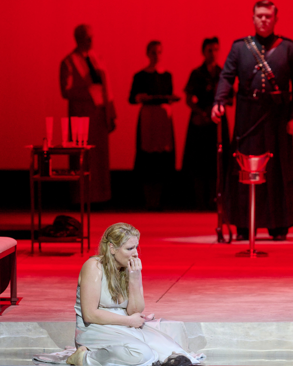 Eine Frau im weißen Kleid sitzt verzweifelt auf dem Boden einer rot ausgeleuchteten Bühne, umgeben von medizinischem Besteck und im Hintergrund stehenden Personen.