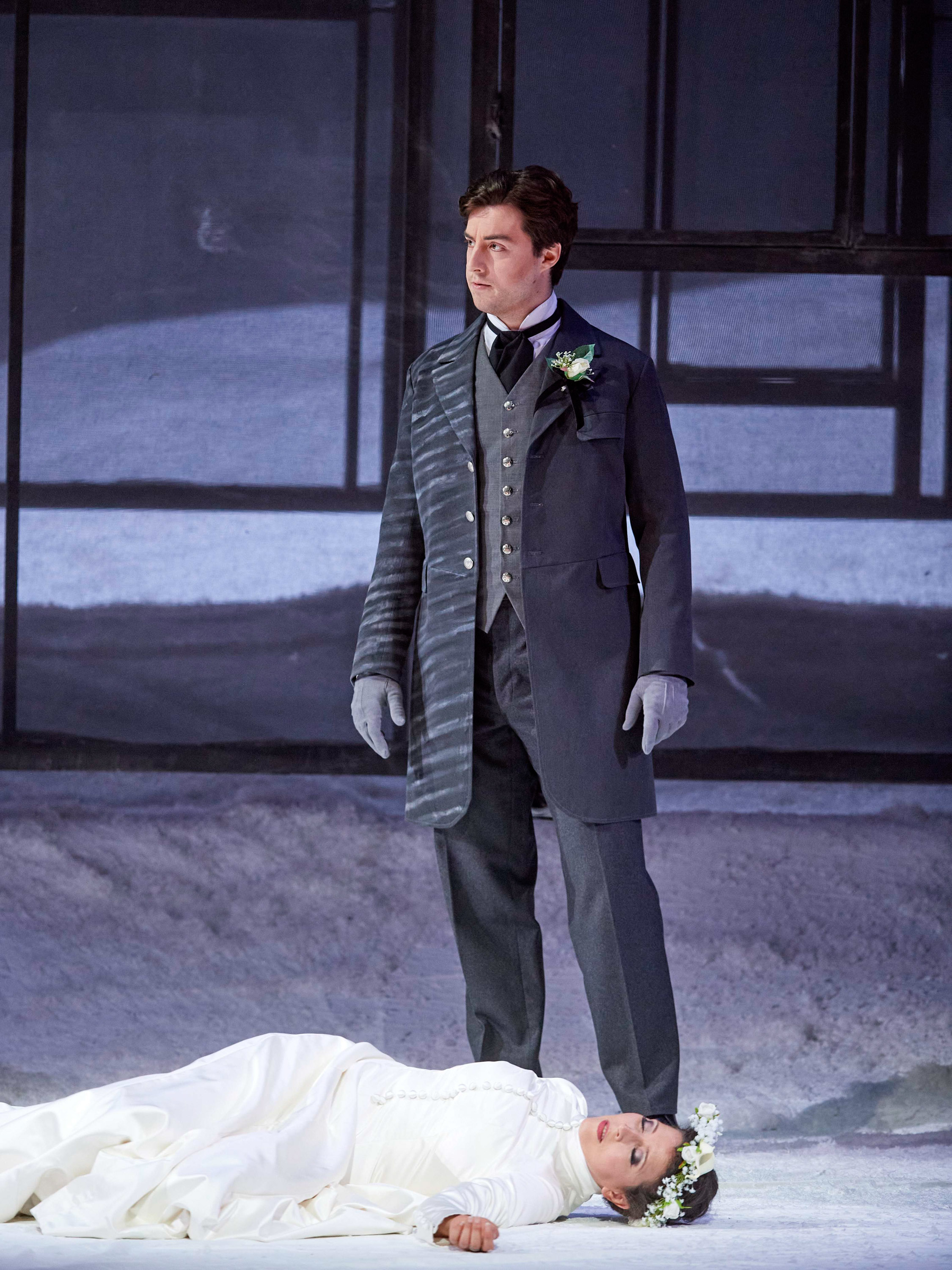 Ein junger Mann in elegantem dunklen Anzug mit grauen Handschuhen steht ernst in einer winterlichen, gläsernen Kulisse.