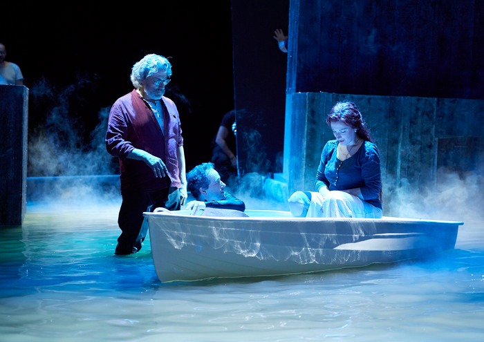 Drei Personen befinden sich in einer nebligen, blau beleuchteten Wasserlandschaft; zwei sitzen in einem kleinen Boot, eine steht daneben.