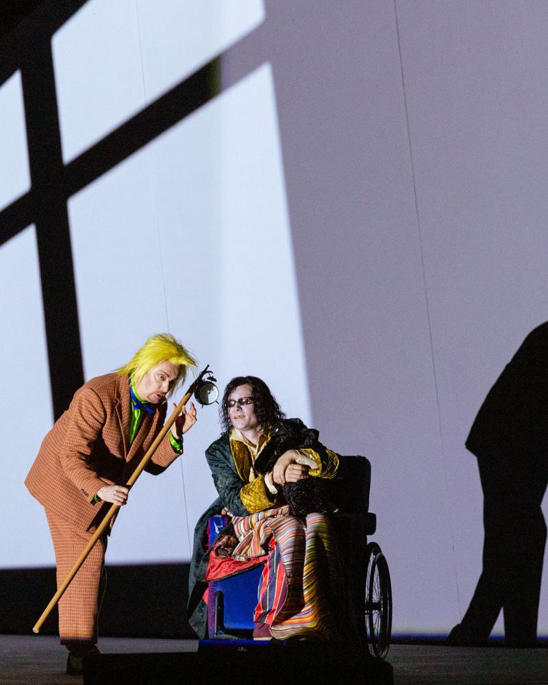 Eine Figur mit gelber Perücke beugt sich zu einer Person im Rollstuhl, beide vor einer Wand mit markanten Schattenprojektionen.