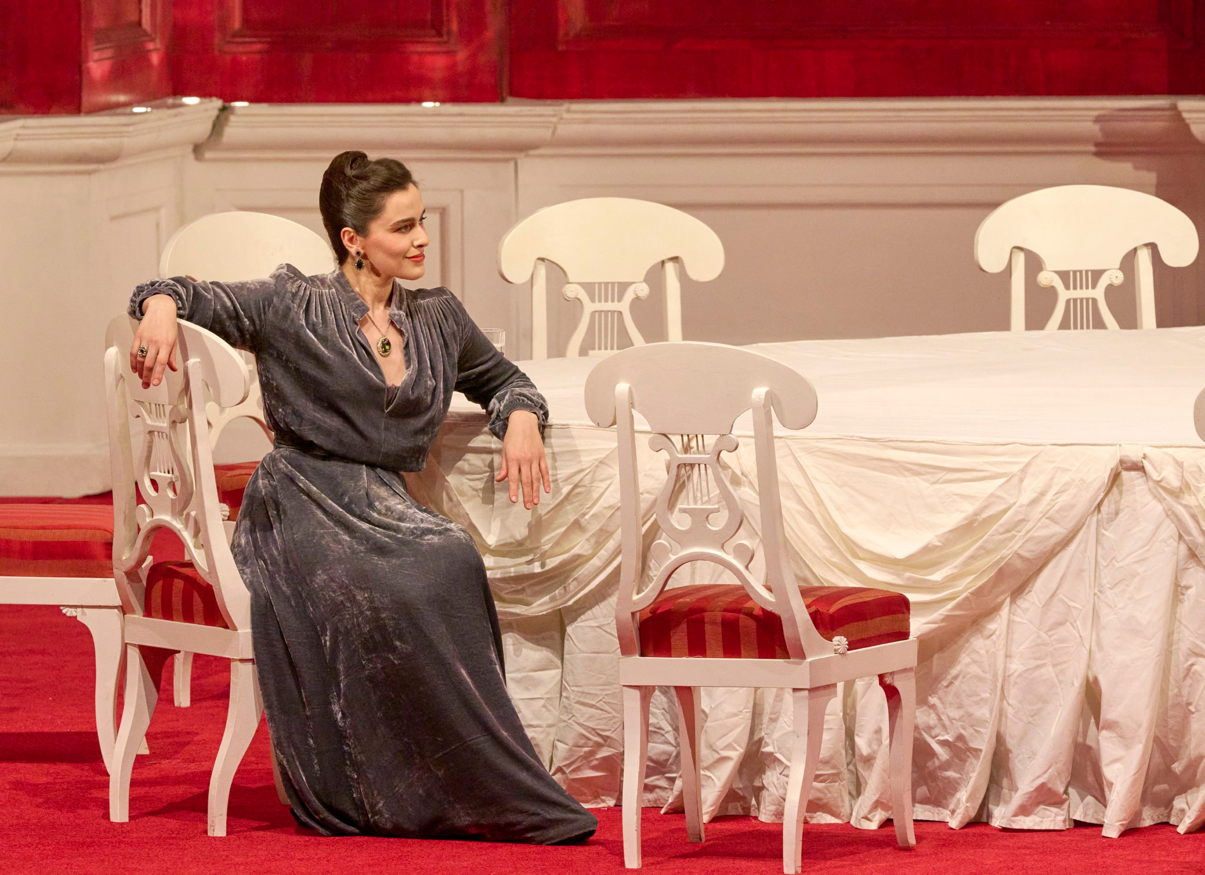 Eine elegant gekleidete Frau sitzt entspannt auf einem Stuhl in einem prunkvoll eingerichteten Raum mit rotem Teppich.