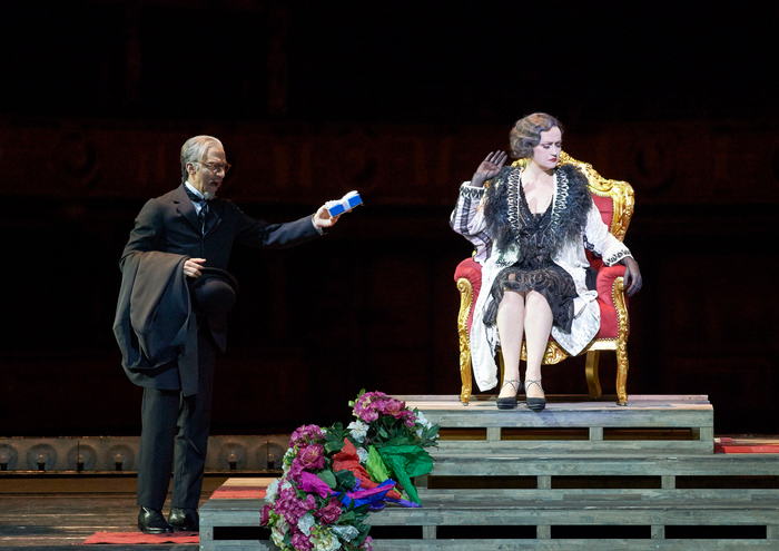 Ein älterer Mann im schwarzen Anzug steht auf einem roten Teppich und zeigt deutet nach links auf eine Treppe, auf der Blumensträuße liegen.