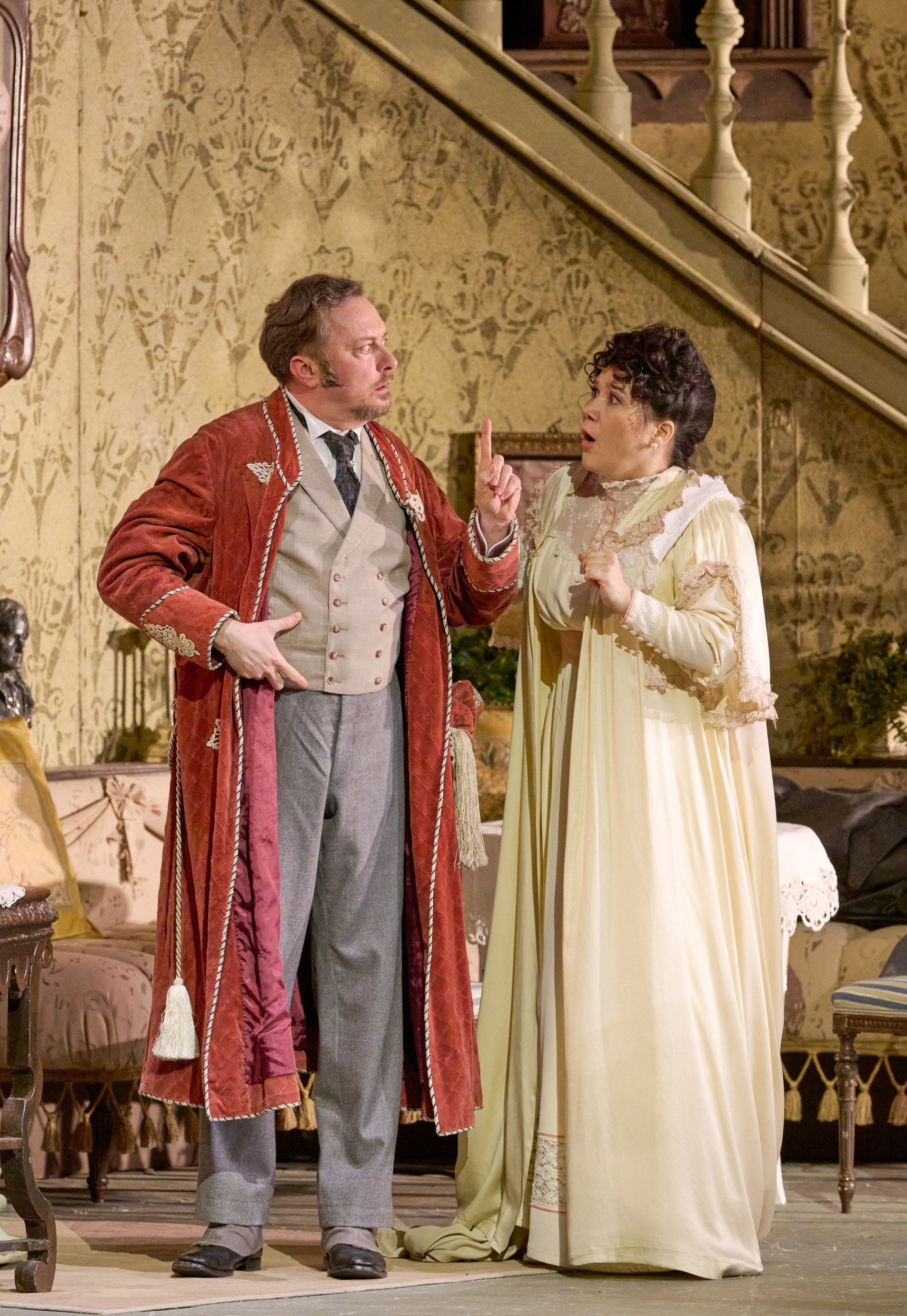 Ein Mann im roten Hausmantel hebt mahnend den Finger, während eine Frau in hellem Nachthemd überrascht reagiert; im Hintergrund eine Treppe mit Tapete.