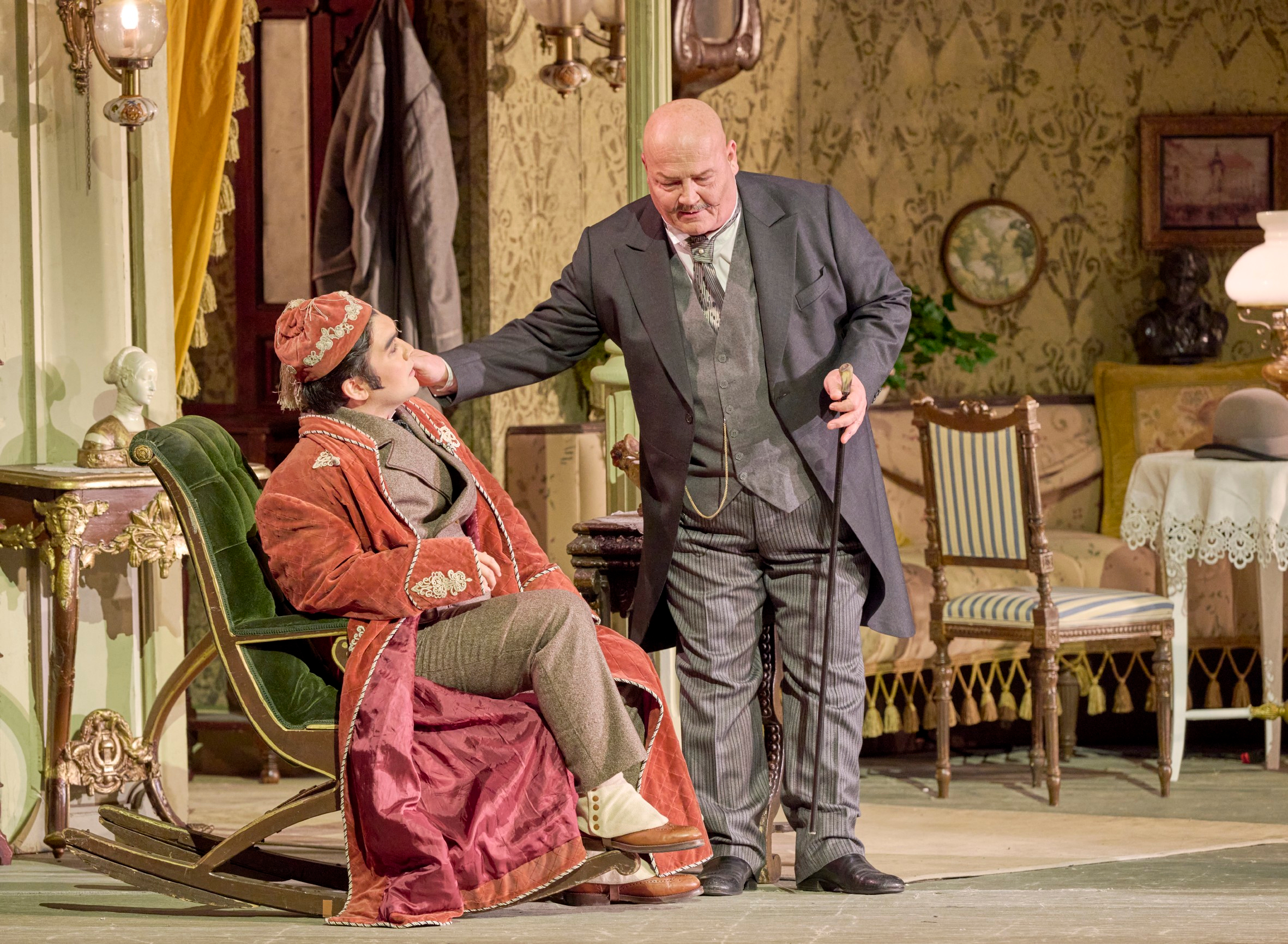 Ein älterer Mann in Anzug spricht beruhigend auf einen sitzenden Mann im roten Morgenmantel ein und berührt sanft dessen Gesicht.